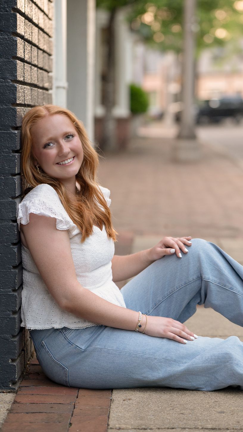 Smiling redhead teen girl in white top and jeans sitting against brick wall for senior portraits.