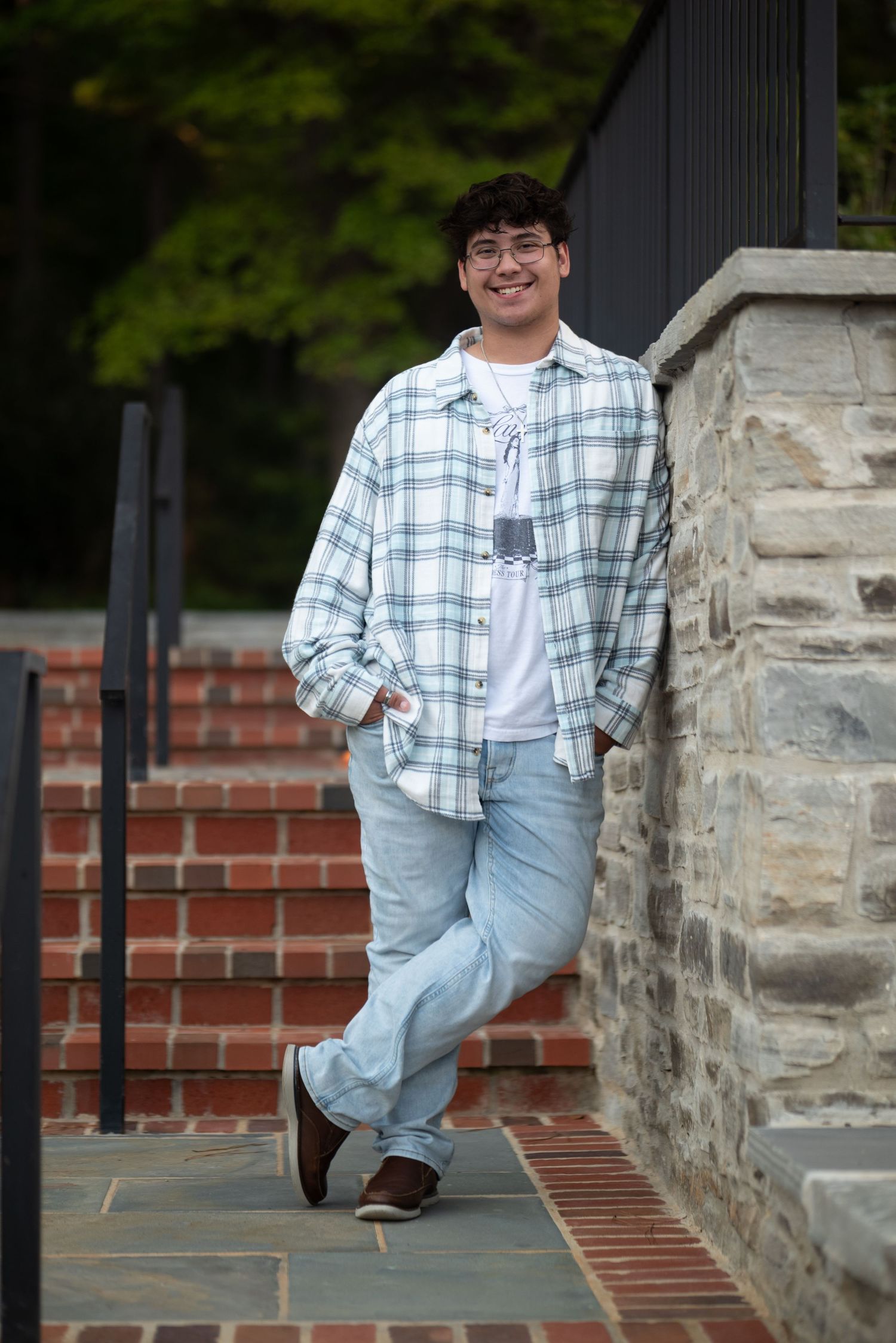 Smiling young man in plaid shirt and jeans leaning against stone wall on brick steps.