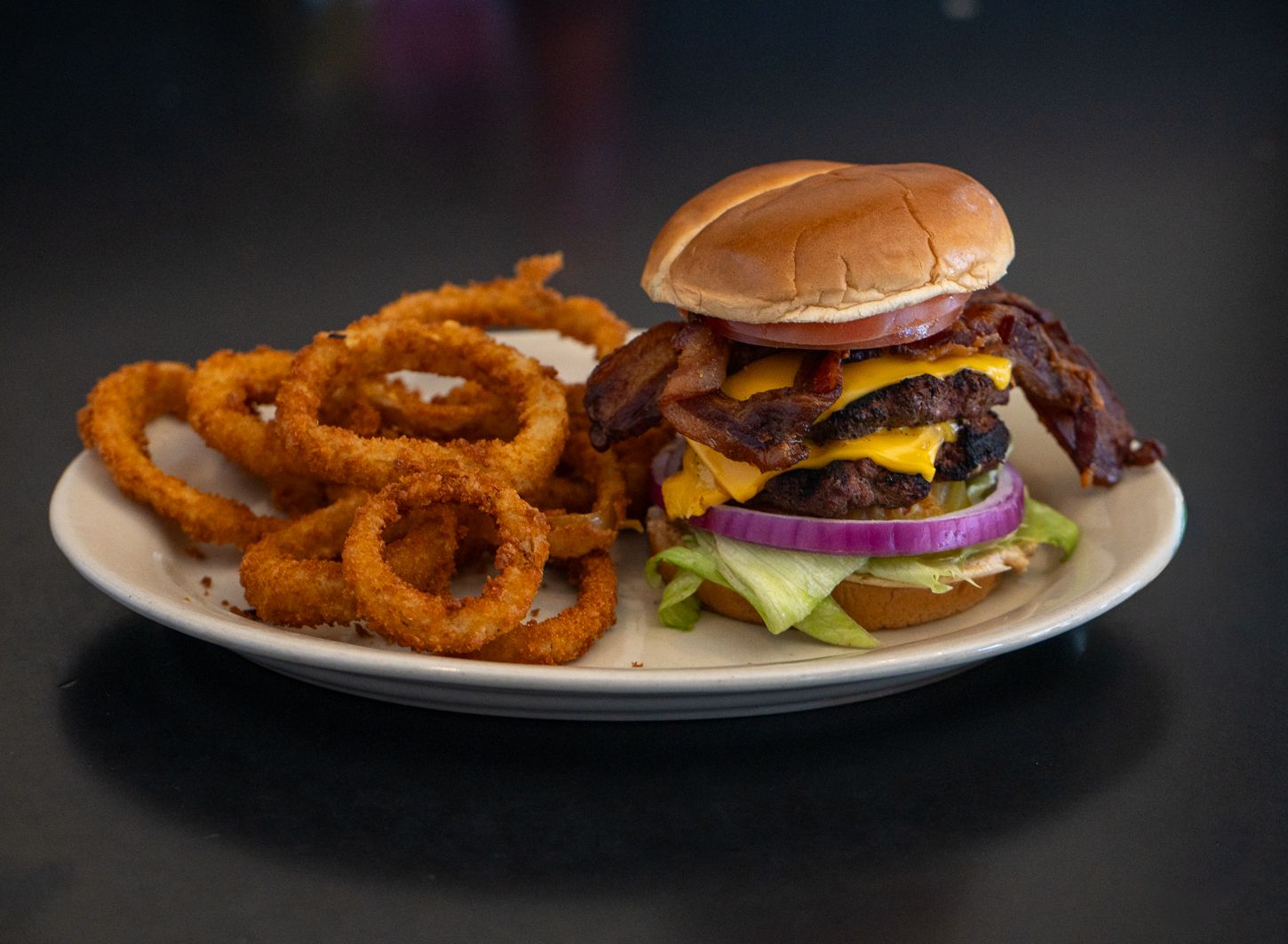 Stacked bacon cheeseburger with lettuce and onion served alongside golden crispy onion rings on a plate.