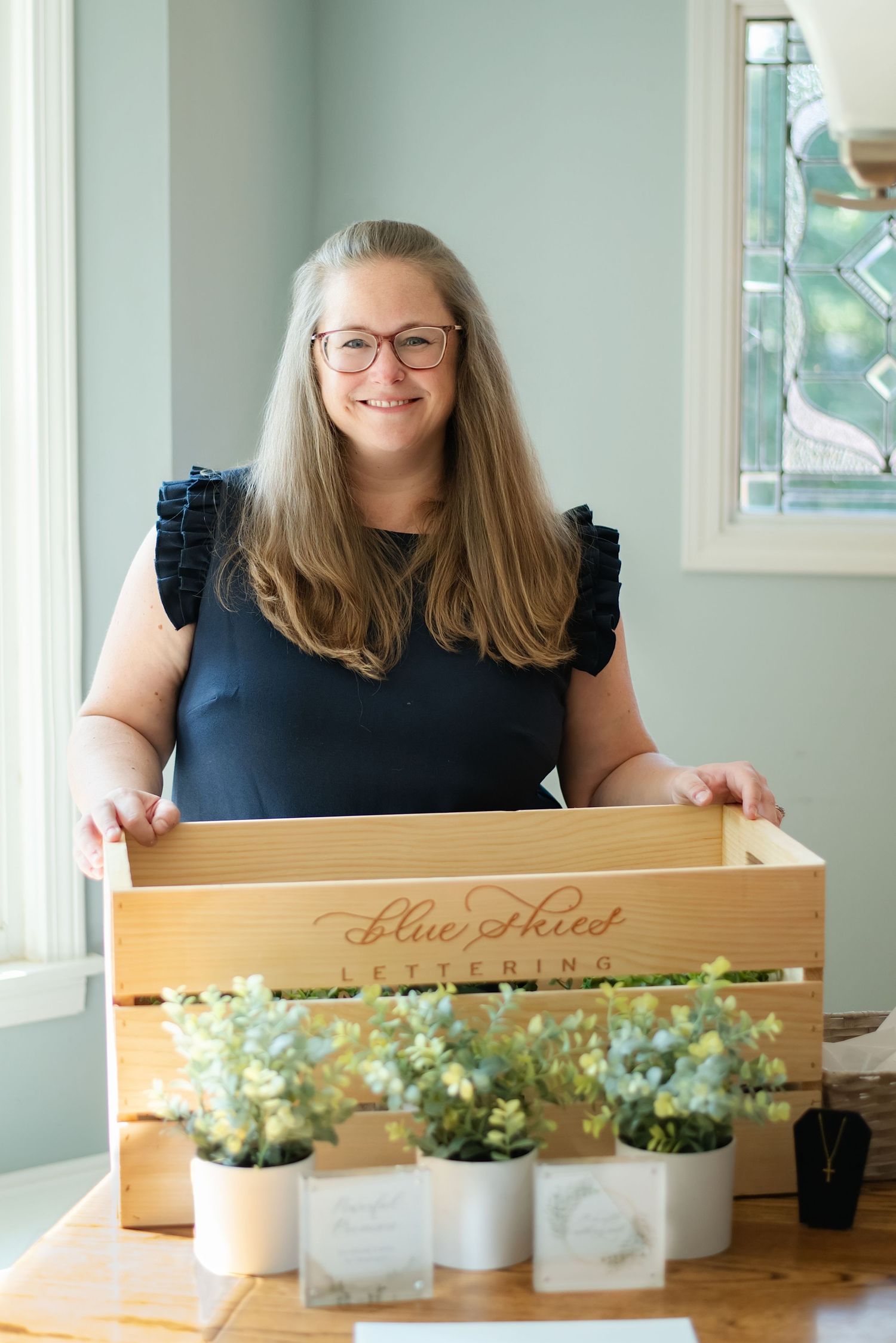 Smiling woman holding a wooden crate labeled Blue Skies Lettering with small potted plants on a table.