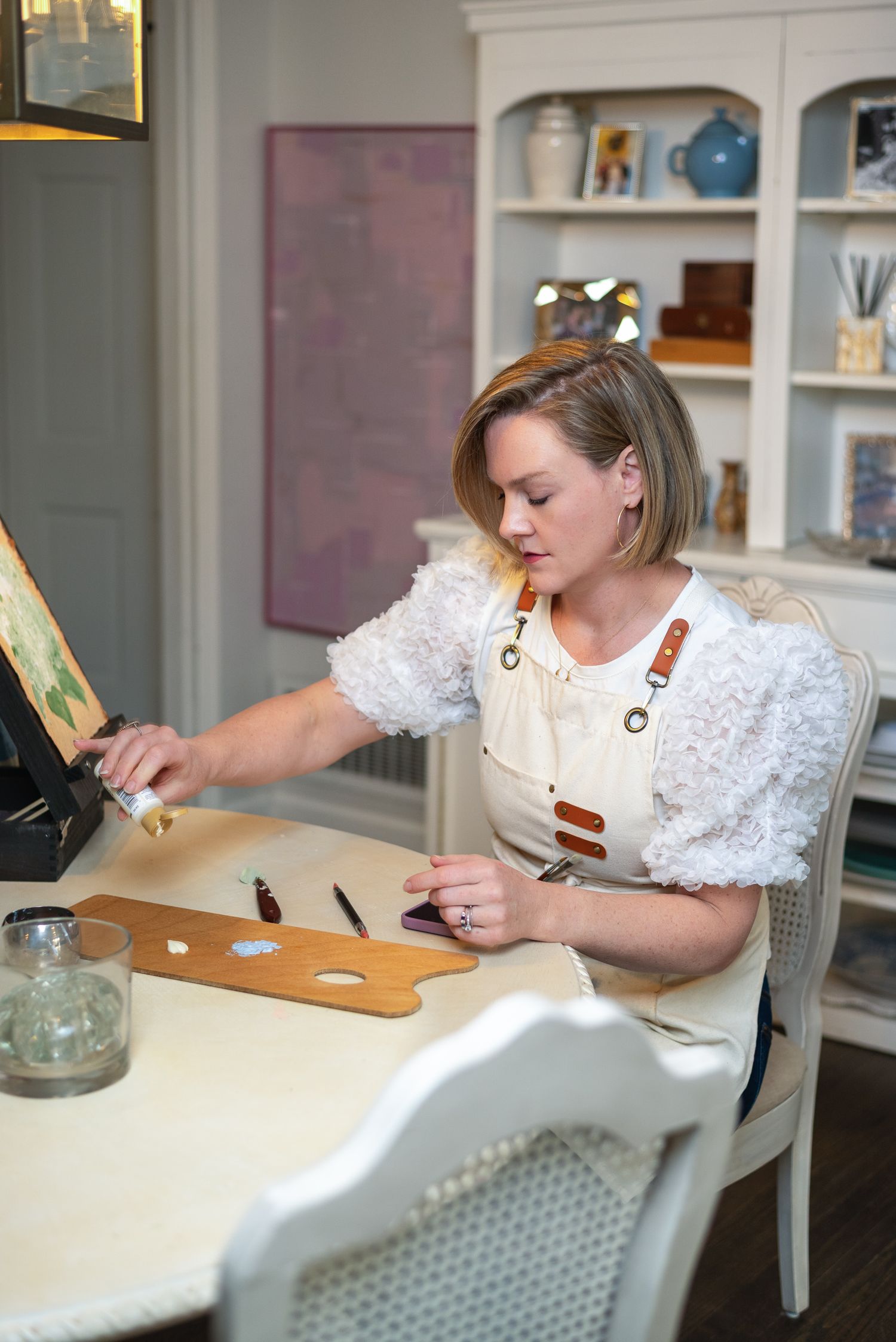 Female artist in white blouse and apron mixing paint on palette at elegant home studio table.