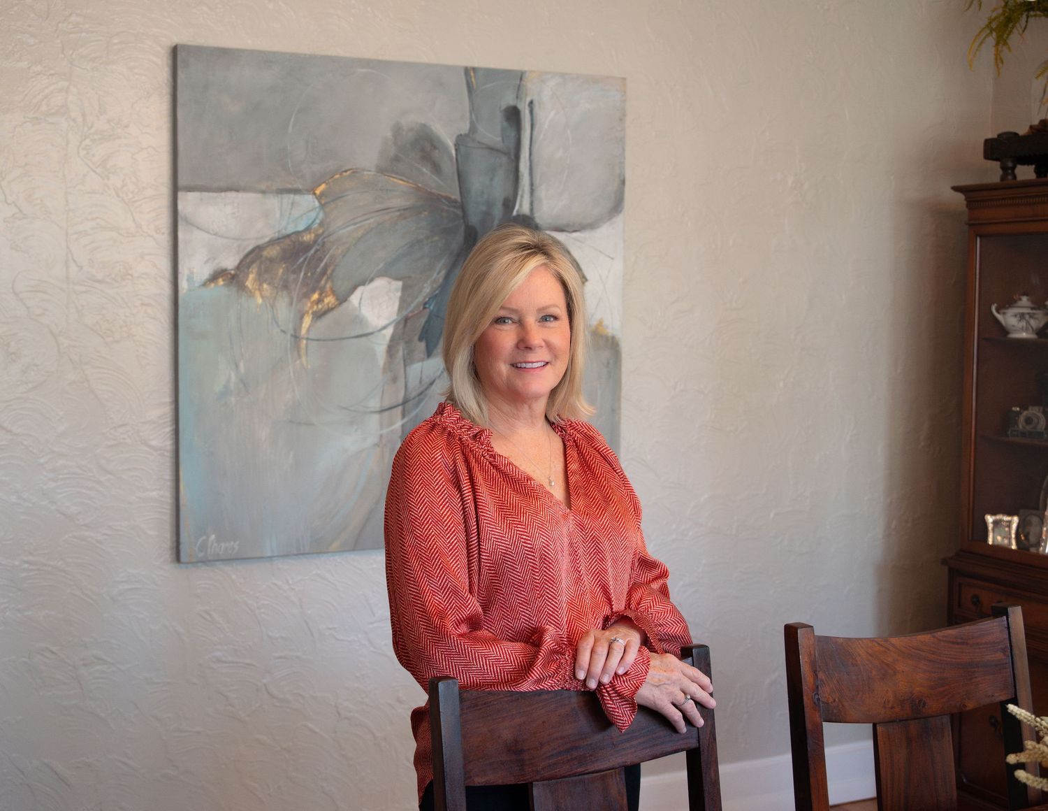 Smiling blonde woman in red top standing at wooden table with abstract painting on wall behind her.