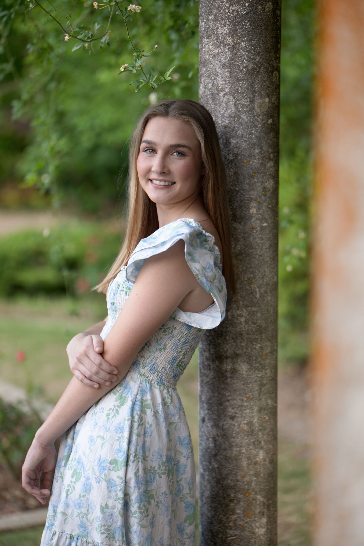 Smiling teenage girl in a floral blue dress leaning against a stone column in a lush garden.