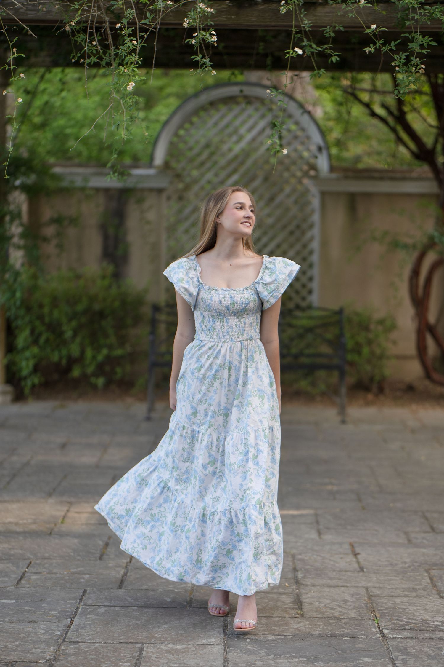 Woman in floral maxi dress with ruffle sleeves posing in garden courtyard with stone path and greenery.
