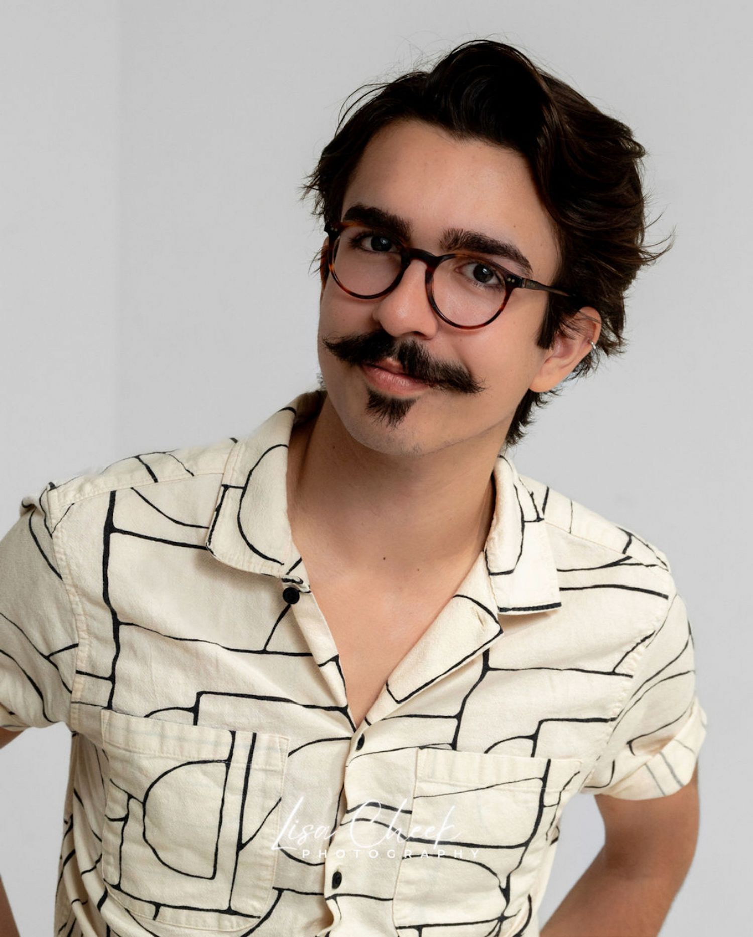 Young man with mustache and glasses wearing a patterned cream shirt against white background.