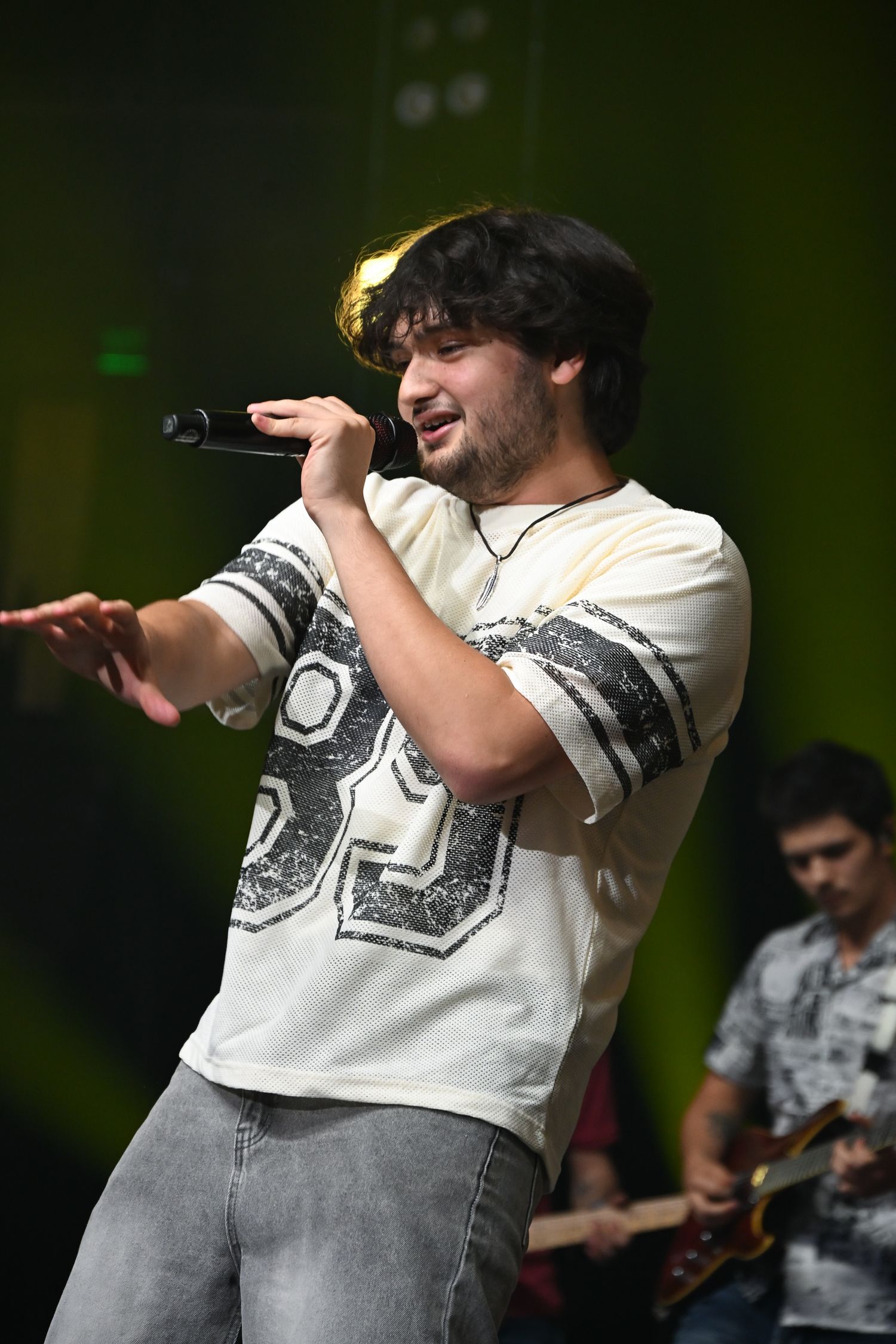 Young male singer performing on stage with microphone, wearing white graphic tee and jeans, guitarist visible in background.
