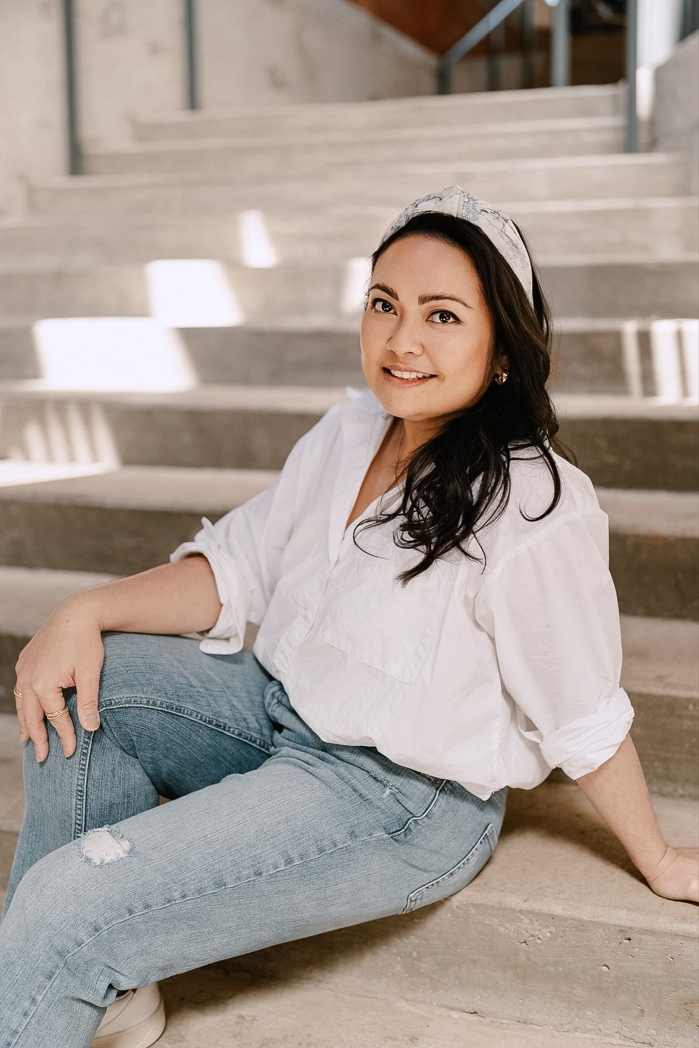 Smiling dark-haired woman in white blouse and jeans sitting on concrete steps outdoors.
