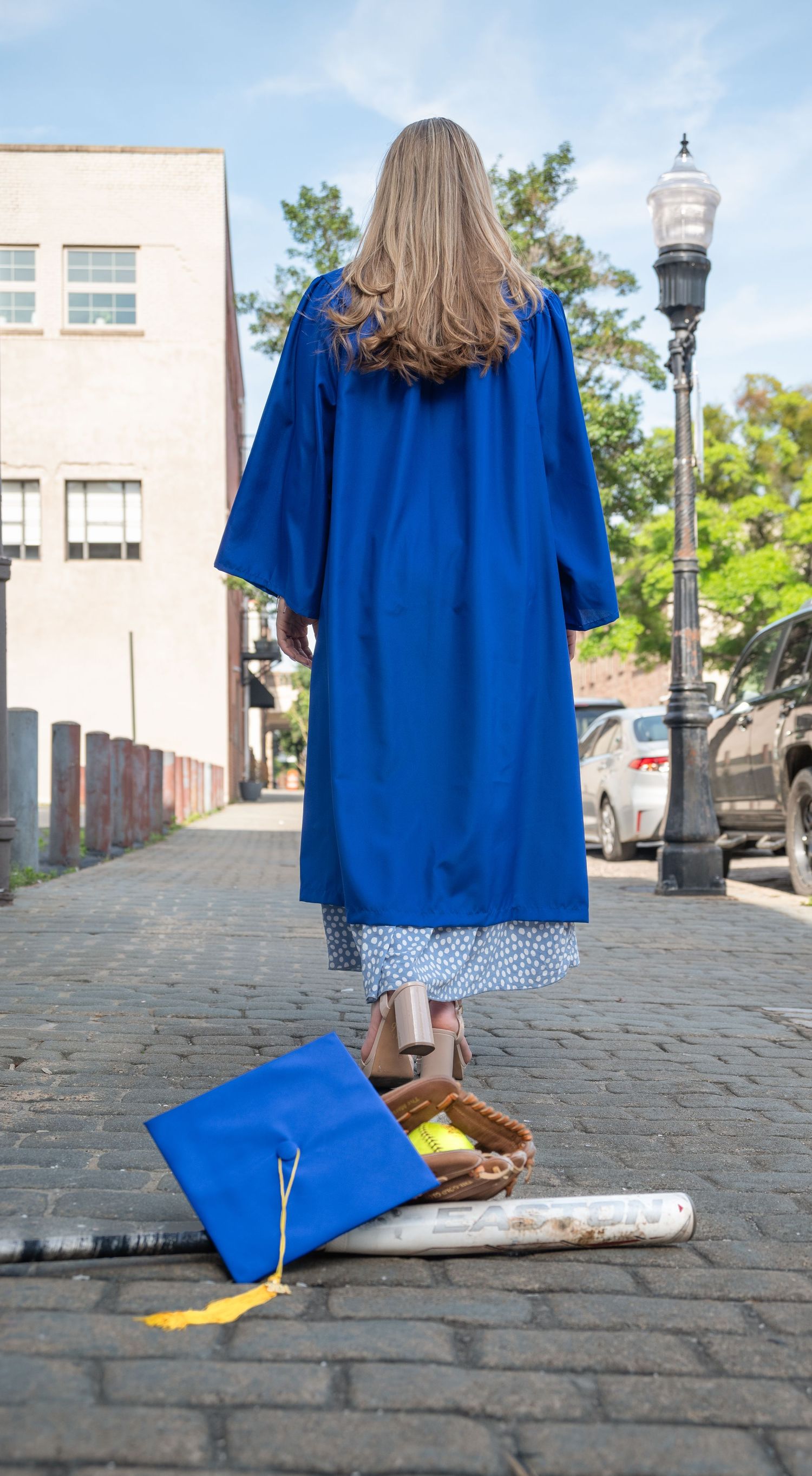 Graduate in blue gown standing on cobblestone street with cap and softball gear on ground behind.