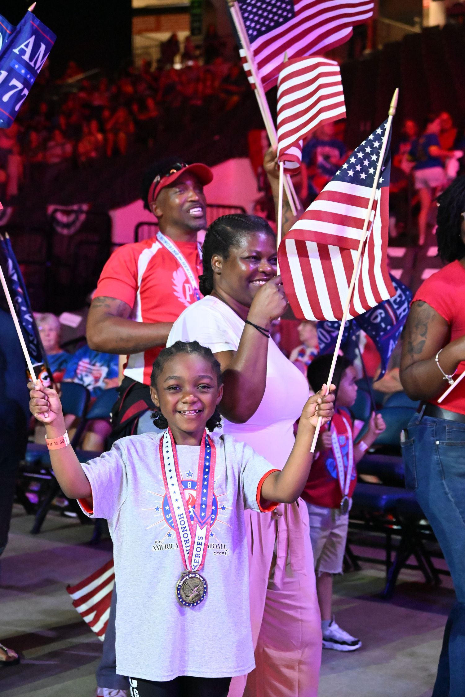 Young girl waves American flag with family at patriotic rally, wearing medal, surrounded by crowd with flags.