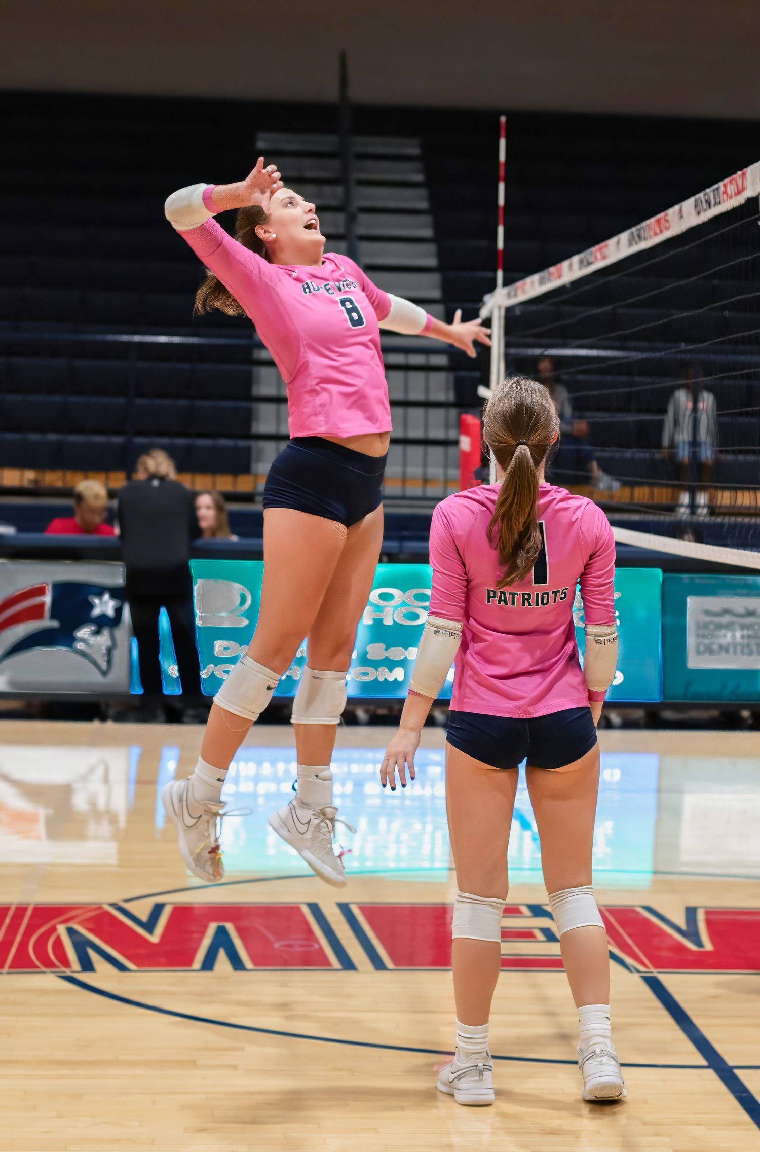 Female volleyball player in pink jersey jumps to spike ball over net as teammate watches during indoor match.
