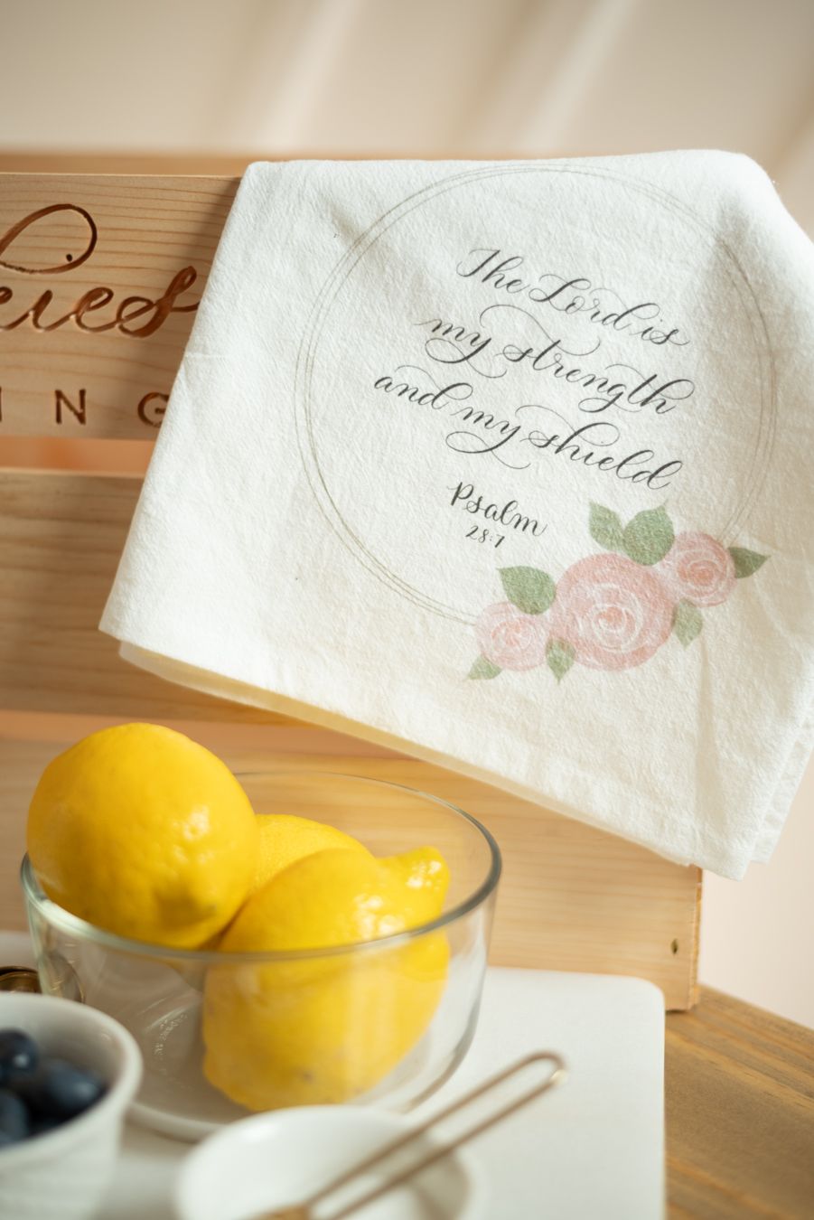 White flour sack towel printed with Psalm 28:7 scripture and floral design displayed beside fresh lemons.