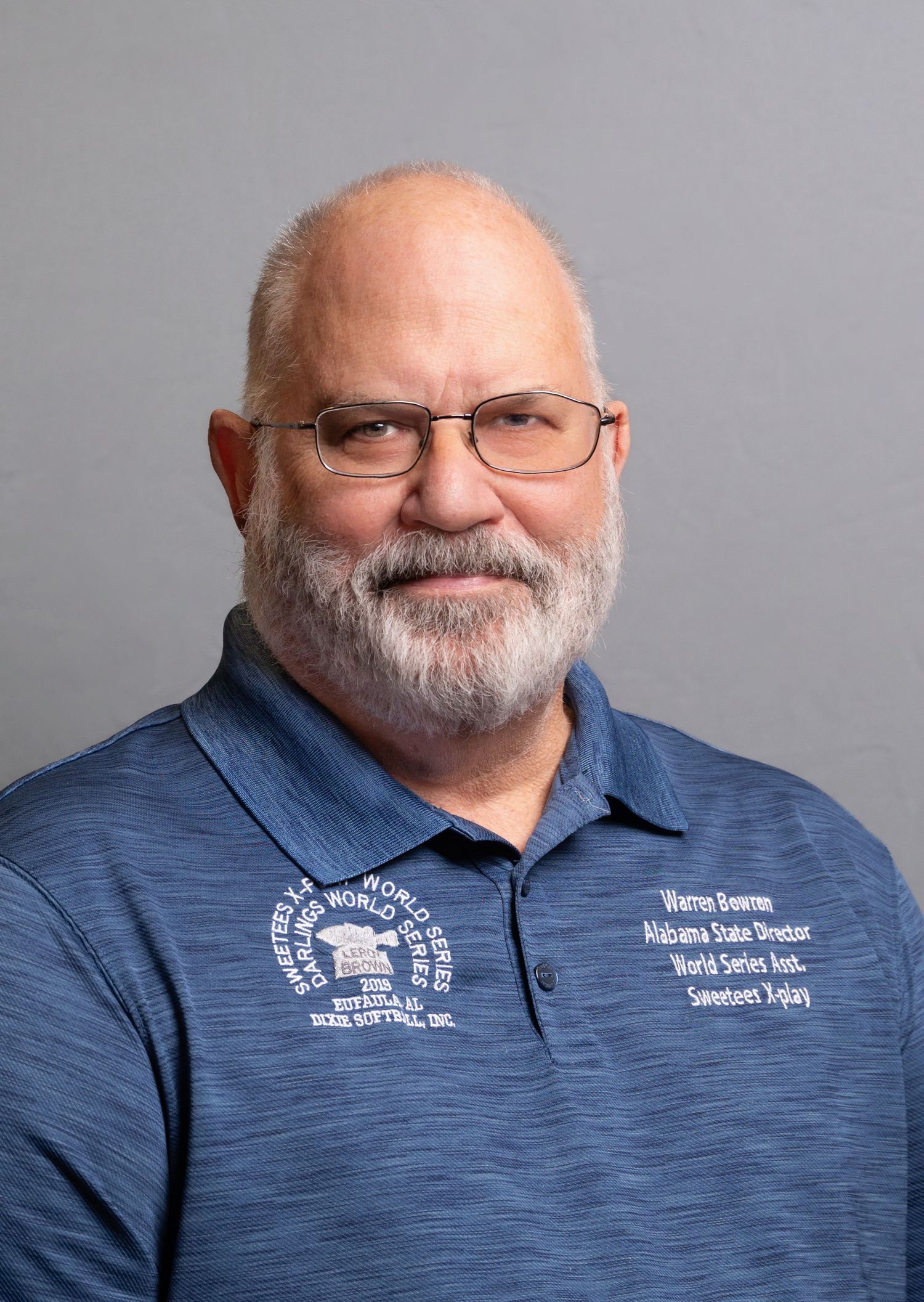 Older man with beard and glasses wearing blue polo shirt in studio portrait.