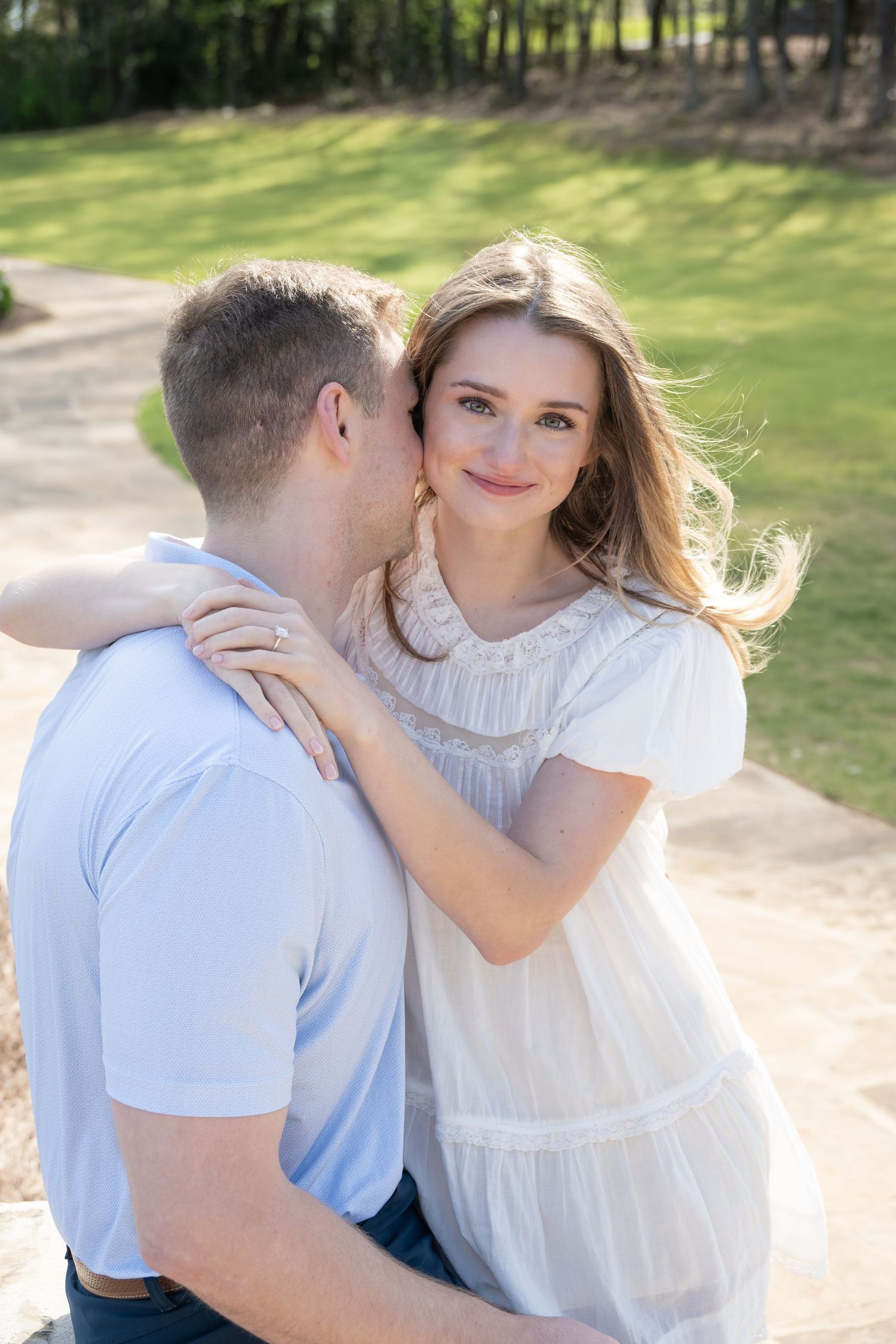 Couple embracing outdoors, woman in white dress smiling as man kisses her cheek.
