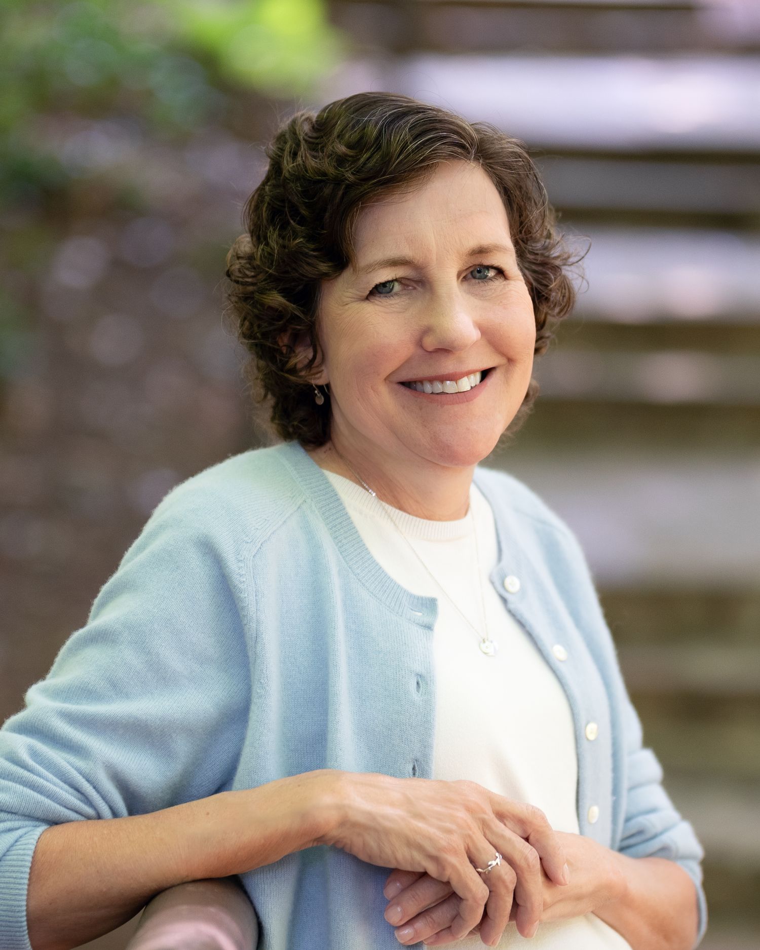 Middle-aged woman in blue cardigan smiling warmly outdoors near stone steps.