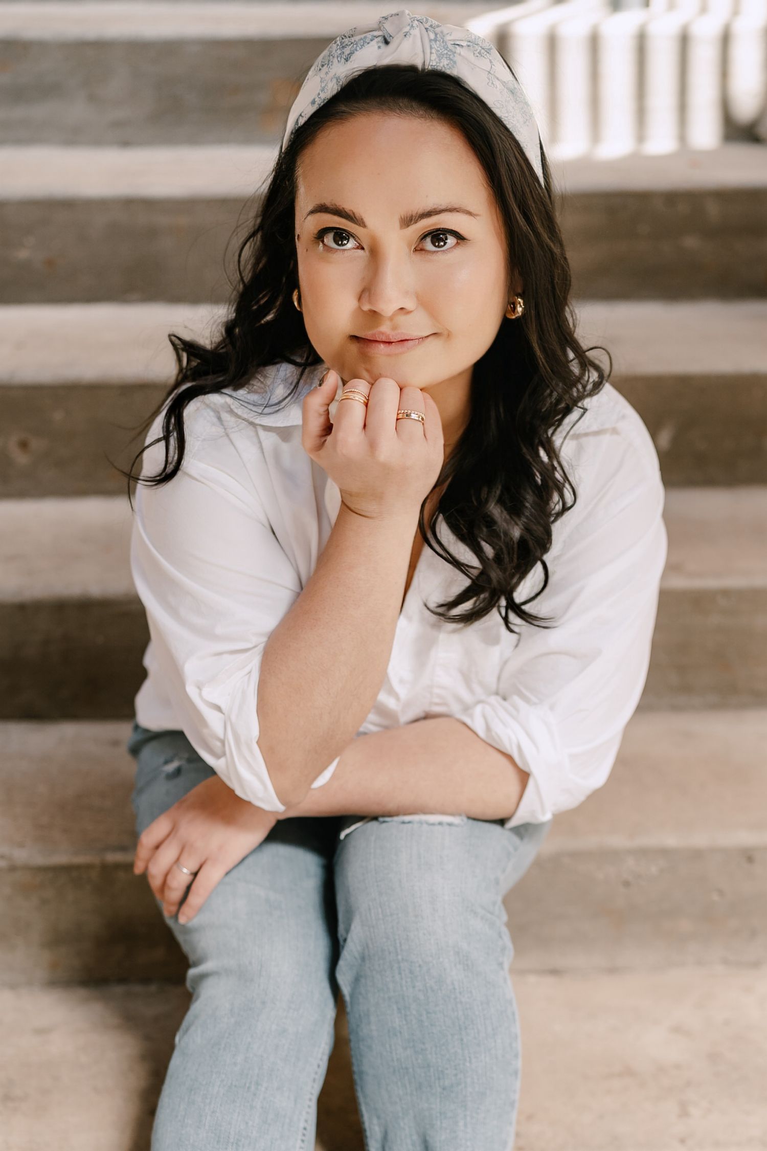 Young woman with dark wavy hair sitting on stairs wearing white shirt and jeans.