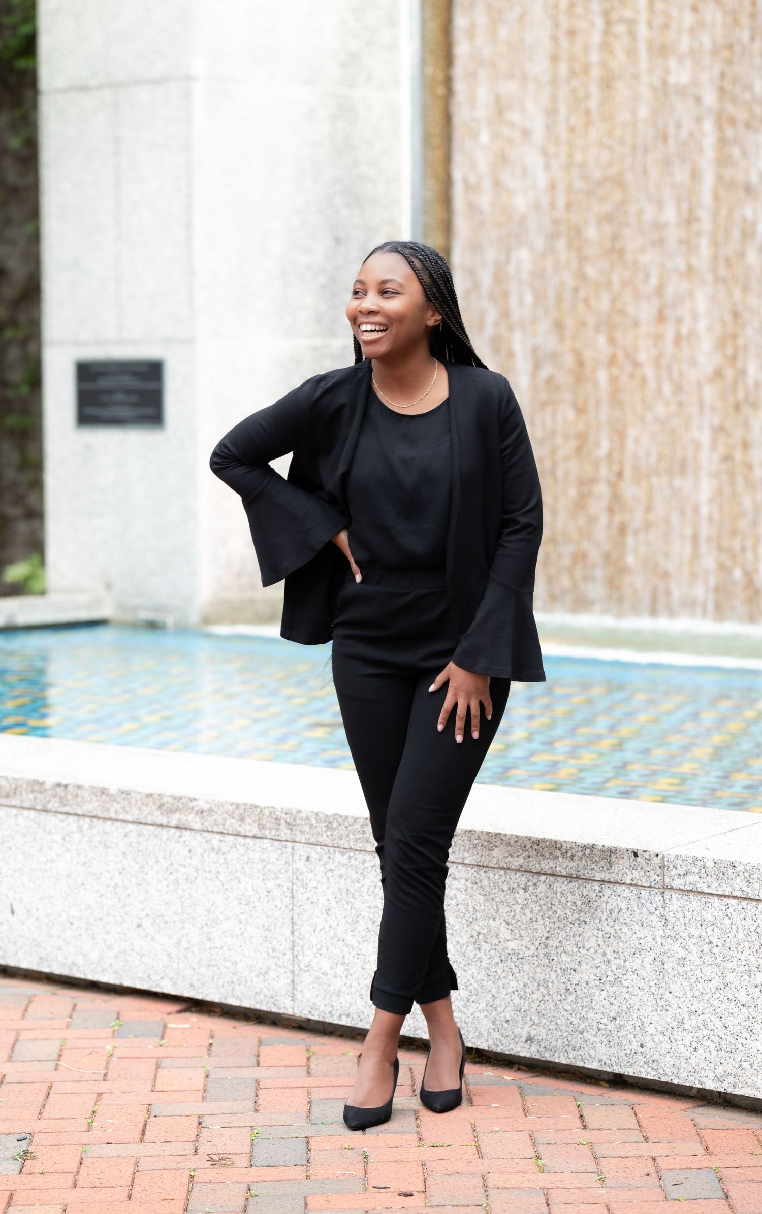 Young Black woman in all-black professional outfit smiling outdoors near a fountain.