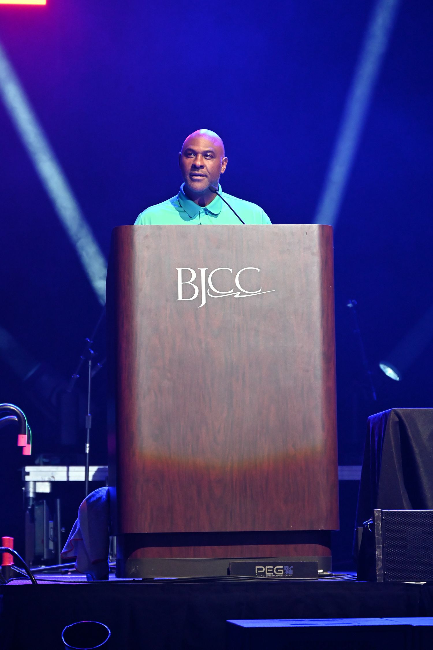 A bald man in a teal shirt speaks at a wooden BJCC podium on a stage with dramatic blue and purple lighting.