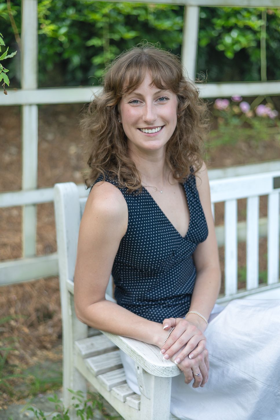 Smiling young woman with curly brown hair sitting on white bench in garden wearing polka dot top.