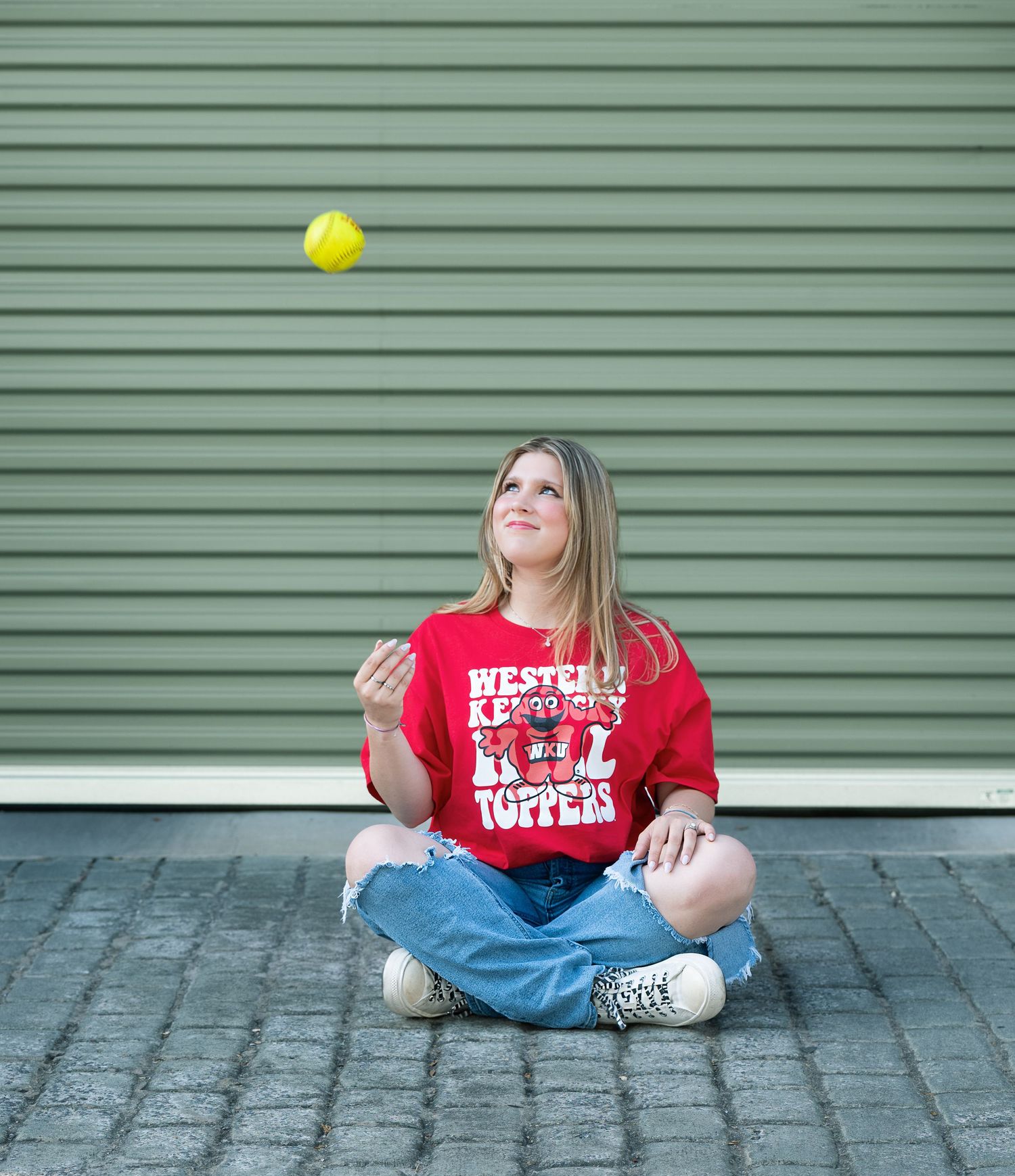 Woman in red Western Kentucky Hilltoppers shirt sits cross-legged on pavement tossing a yellow softball in the air.
