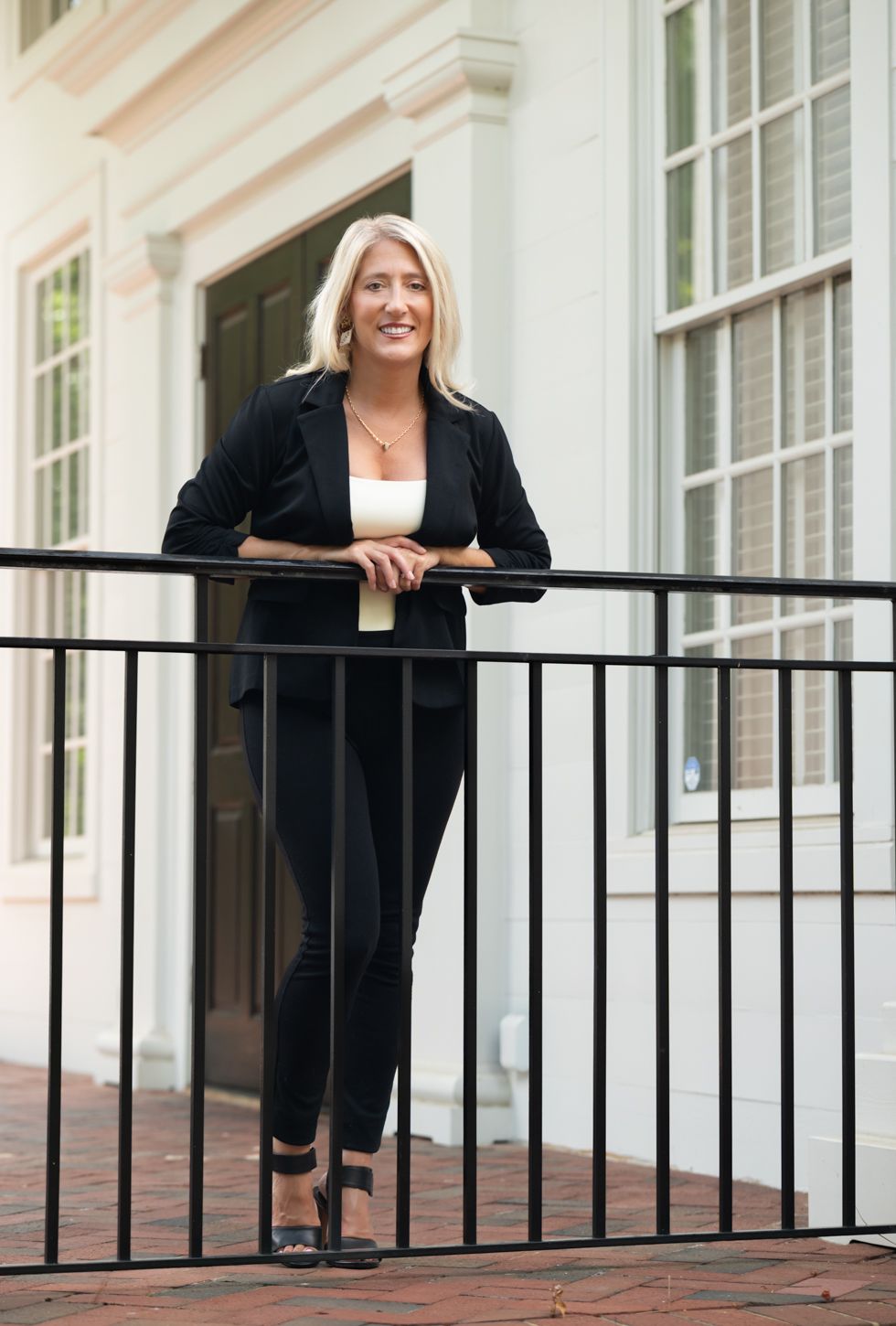 Blonde woman in black blazer and pants leaning on a railing outside a white building.
