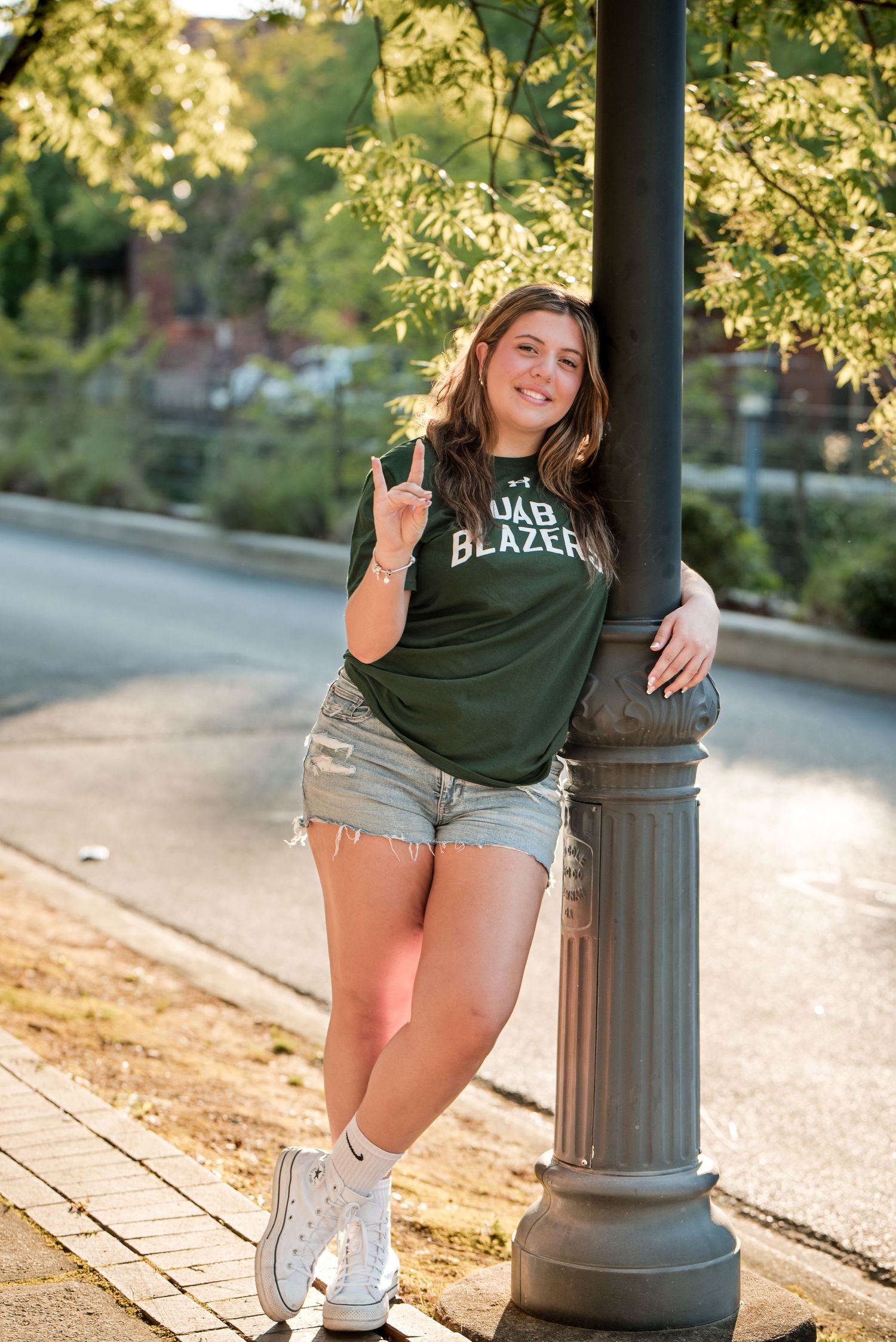 Teen girl in UAB Blazers shirt posing by lamppost on sunny street.