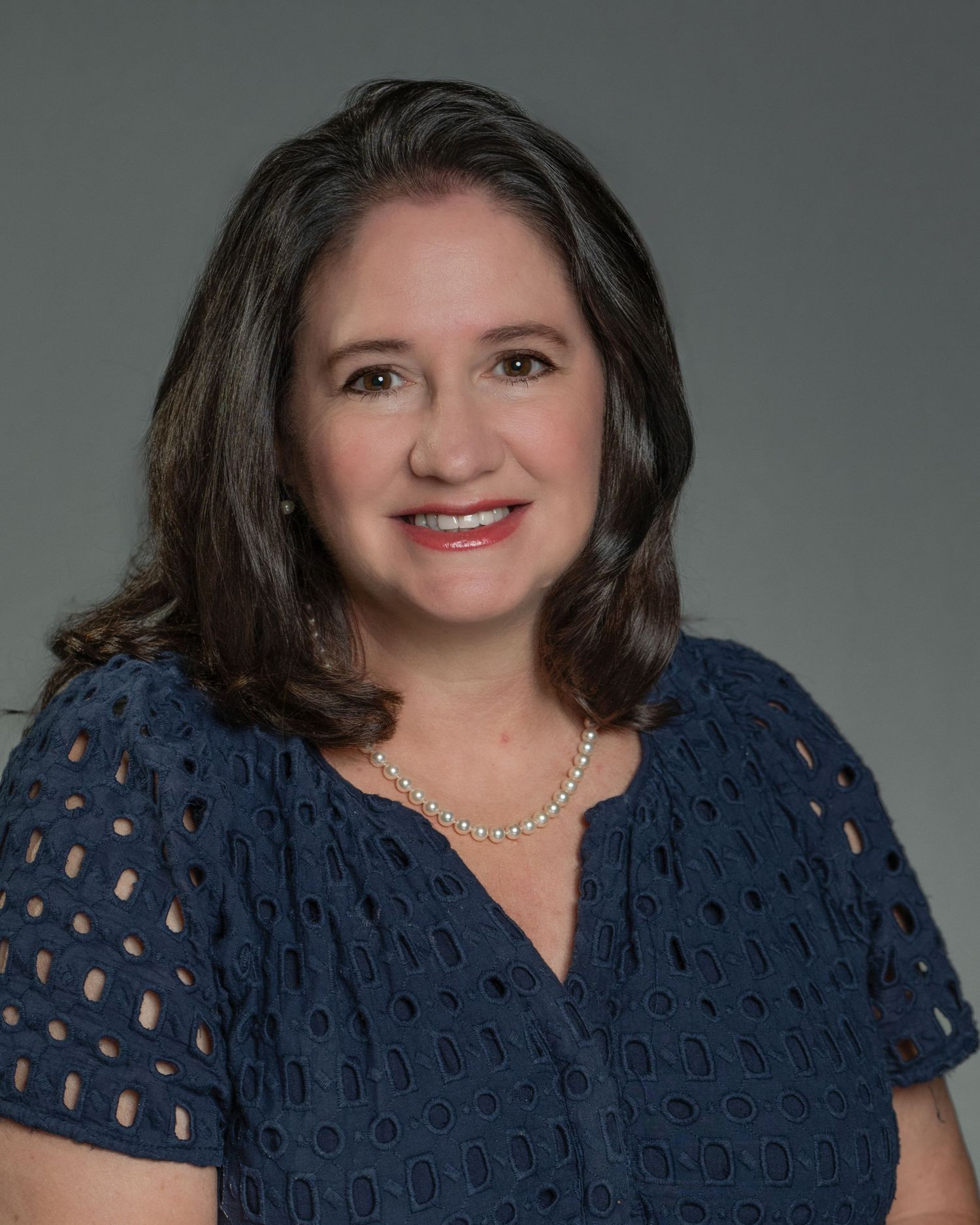 Smiling woman with dark hair wearing navy blue eyelet top and pearl necklace for professional headshot.