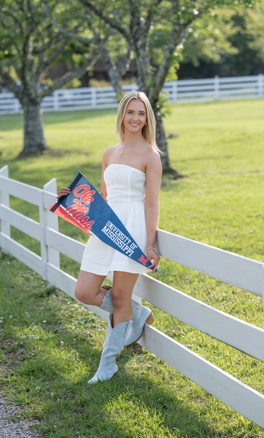 Smiling blonde woman in white dress and gray boots holding University of Mississippi Ole Miss pennant by white fence.