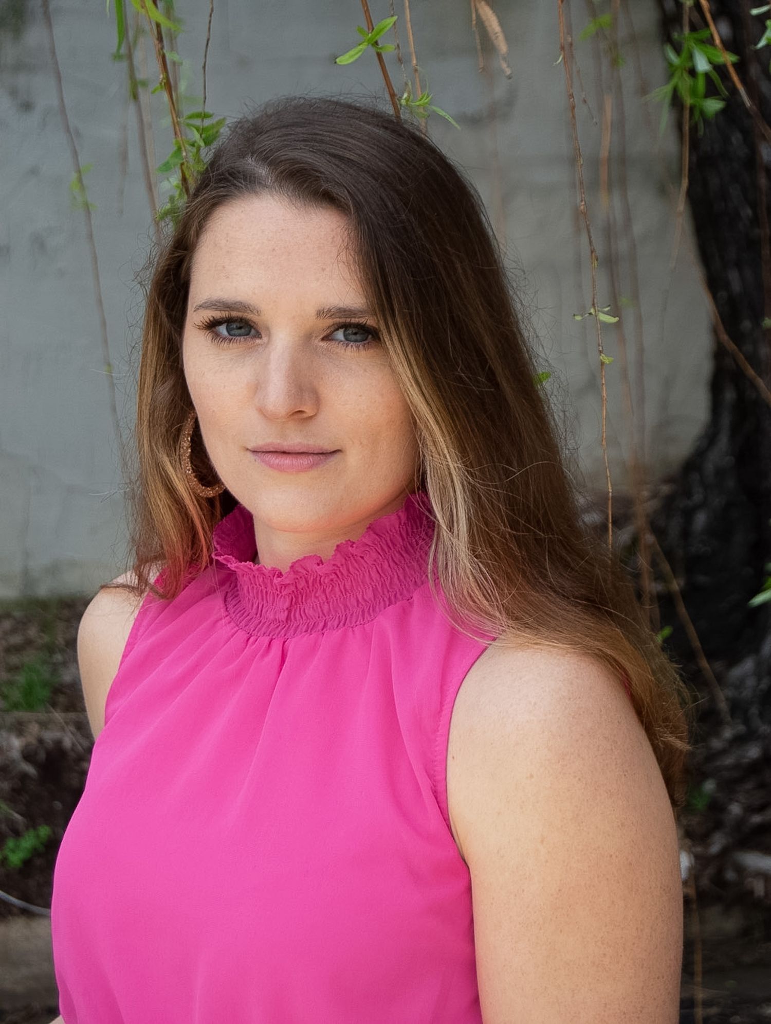 Woman with brown hair in a pink sleeveless top posing outdoors near hanging branches.