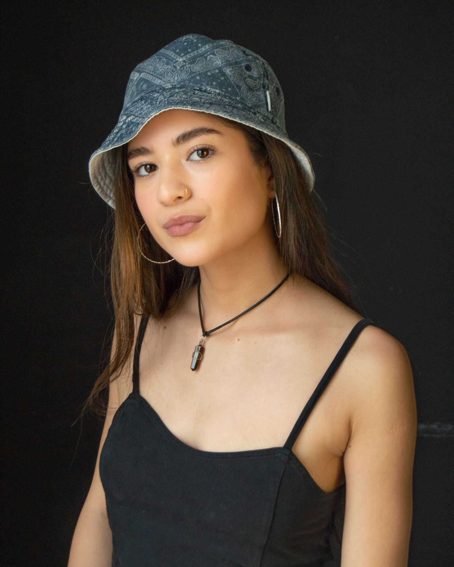 Young woman wearing denim bucket hat and black top poses for artistic portrait on dark background.