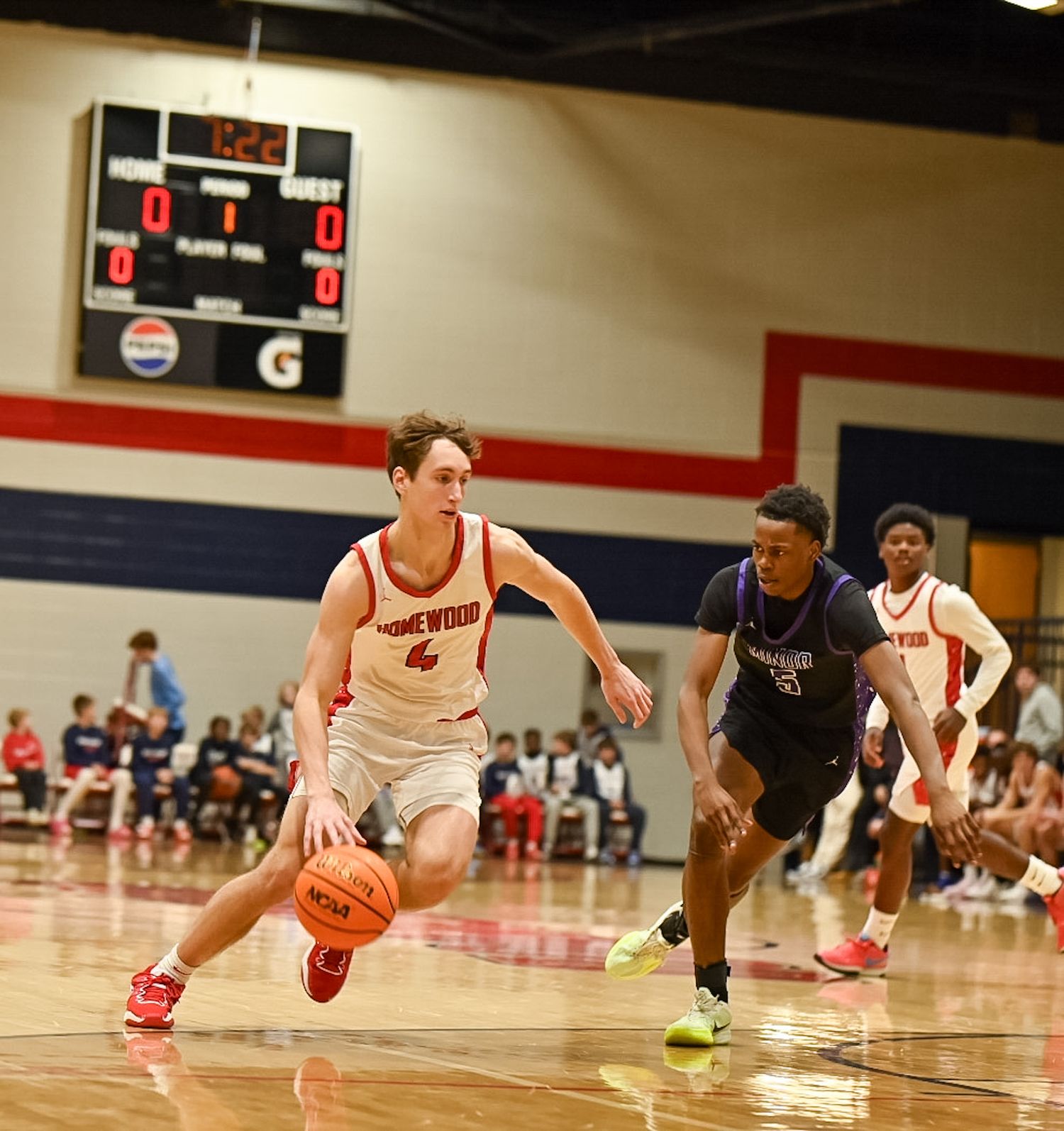 High school basketball player in white Homewood jersey dribbles past defender during indoor game.