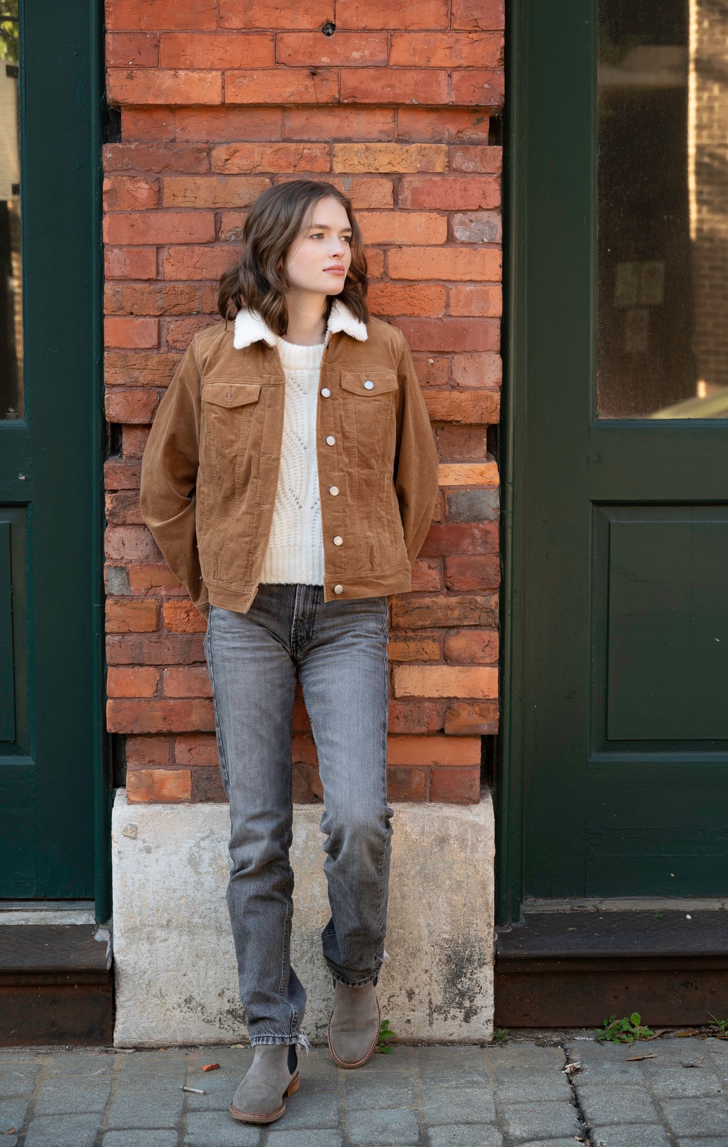 Woman in brown corduroy sherpa jacket, cream sweater, gray jeans, and tan ankle boots leaning against brick wall.