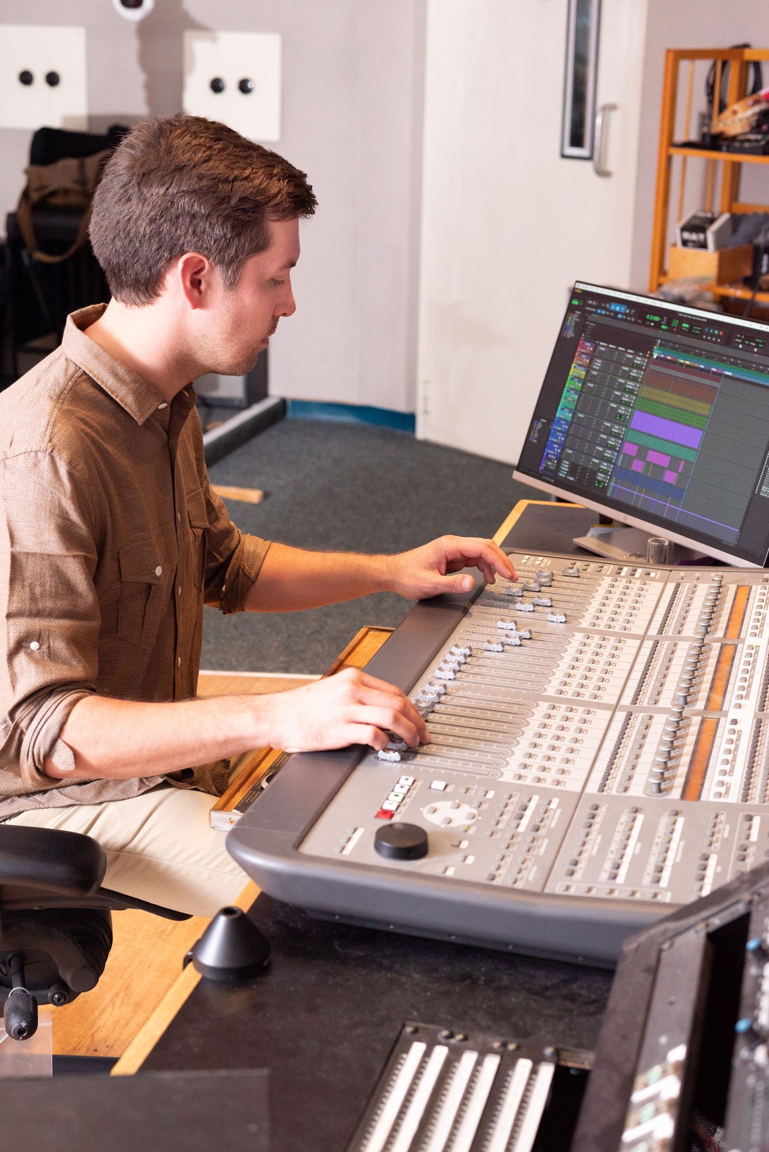 Young man operating a professional audio mixing console in a recording studio with a computer monitor.