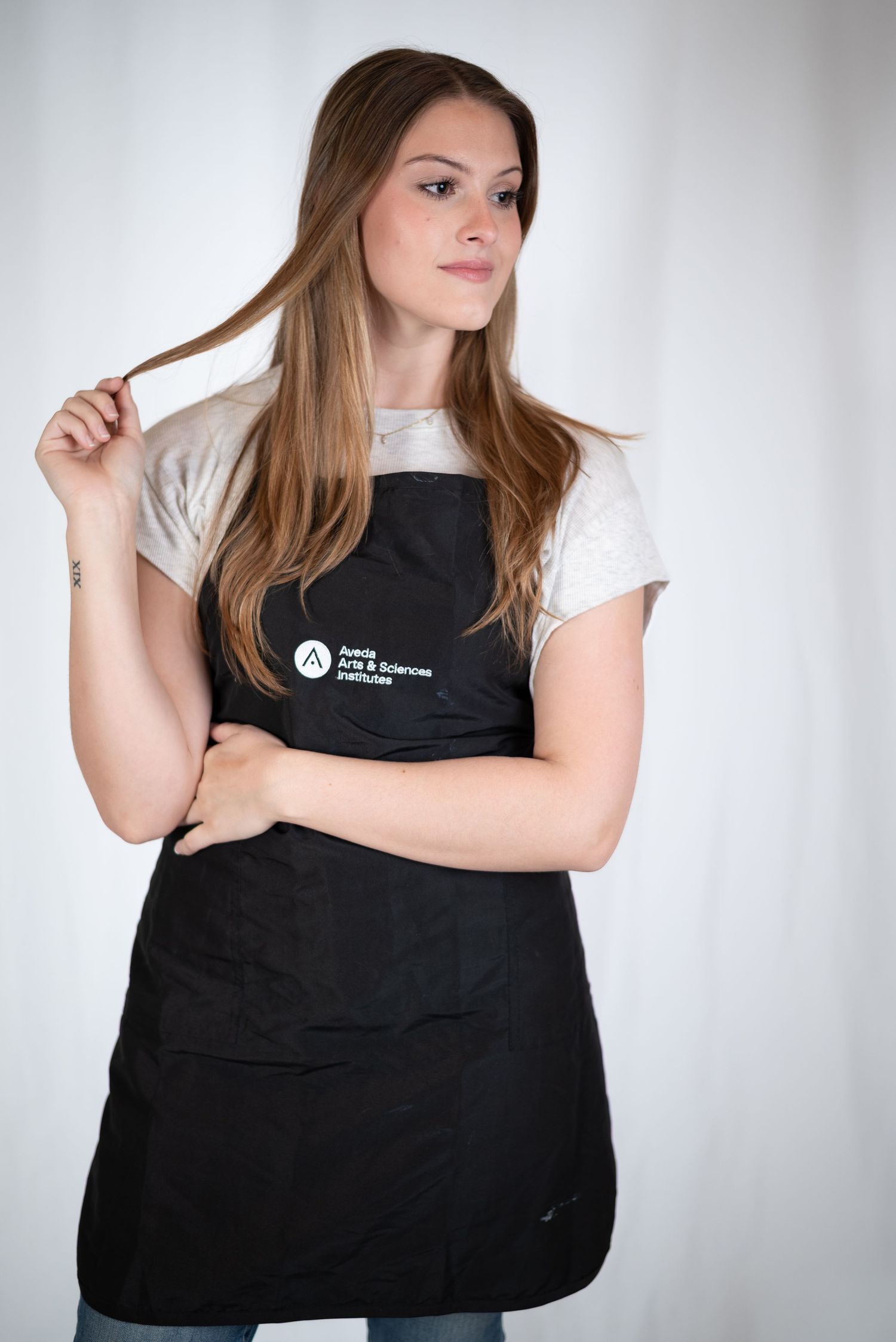 Young woman in black apron from Aveda looks away from camera holding a strand of hair.