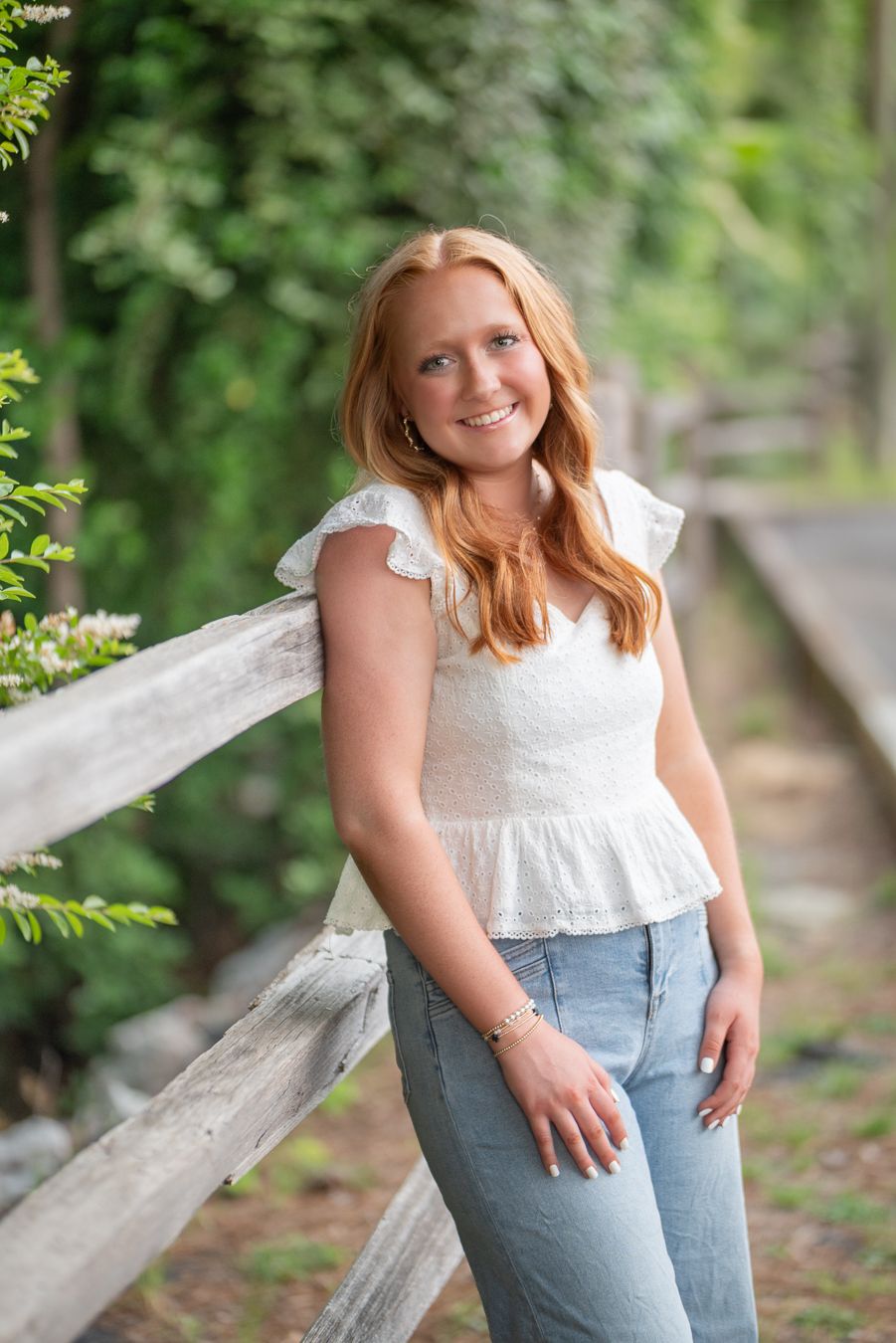 Smiling teenage girl with red hair in white ruffled top and jeans leaning on wooden fence outdoors.