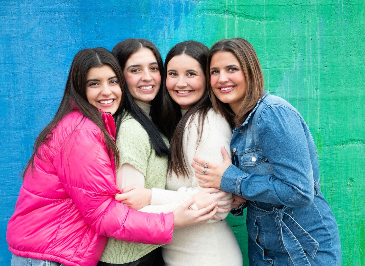 Four smiling women hugging together in front of a colorful blue and green painted wall.
