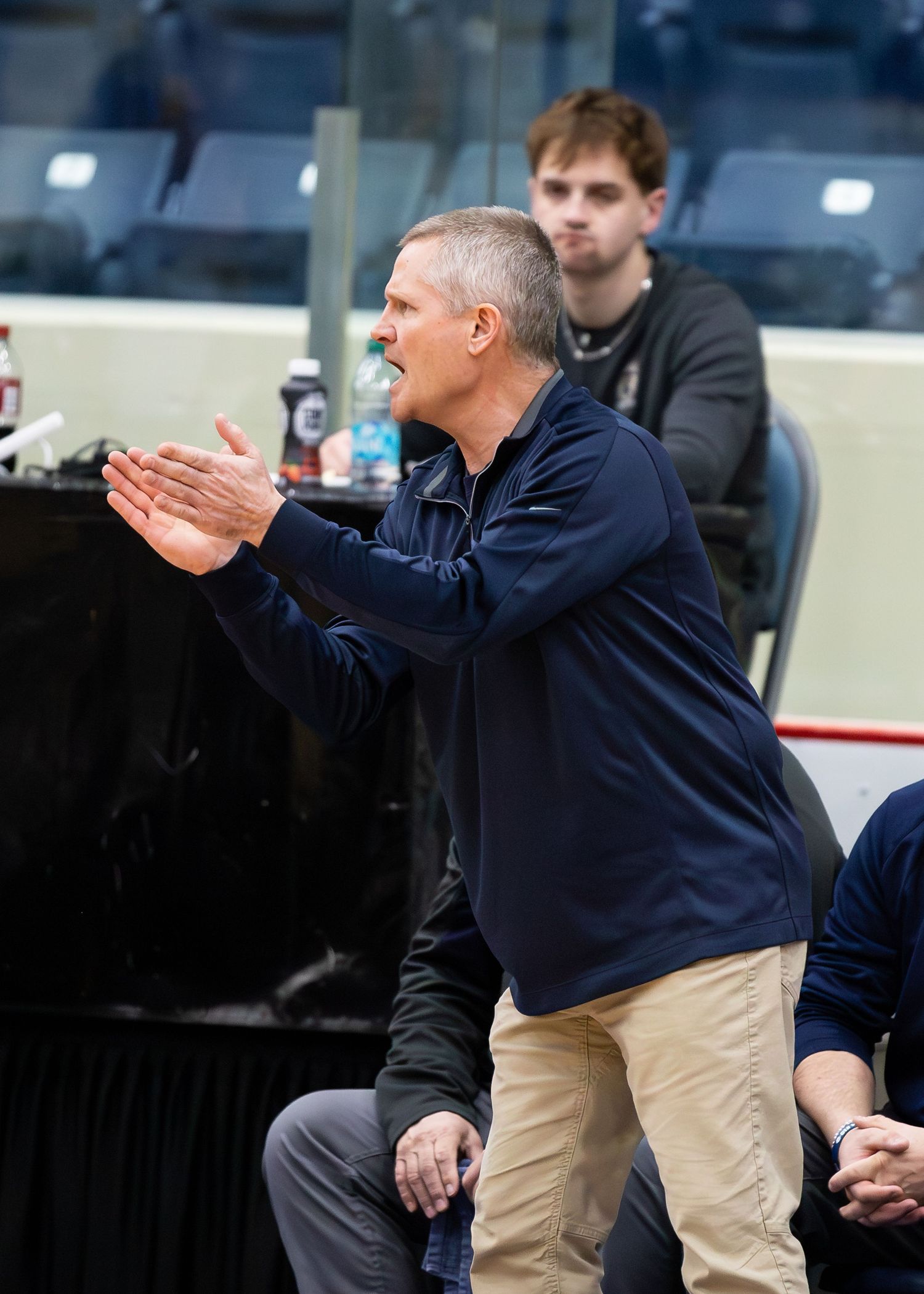 Basketball coach in navy jacket claps encouragingly from sideline during high school game.