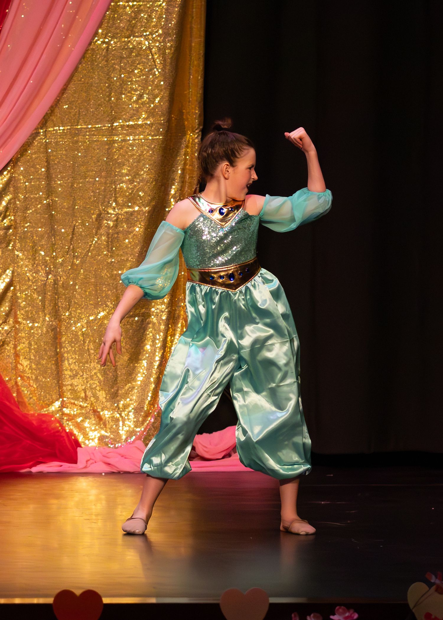 Young dancer in teal Jasmine costume performs on stage with gold sequin backdrop.