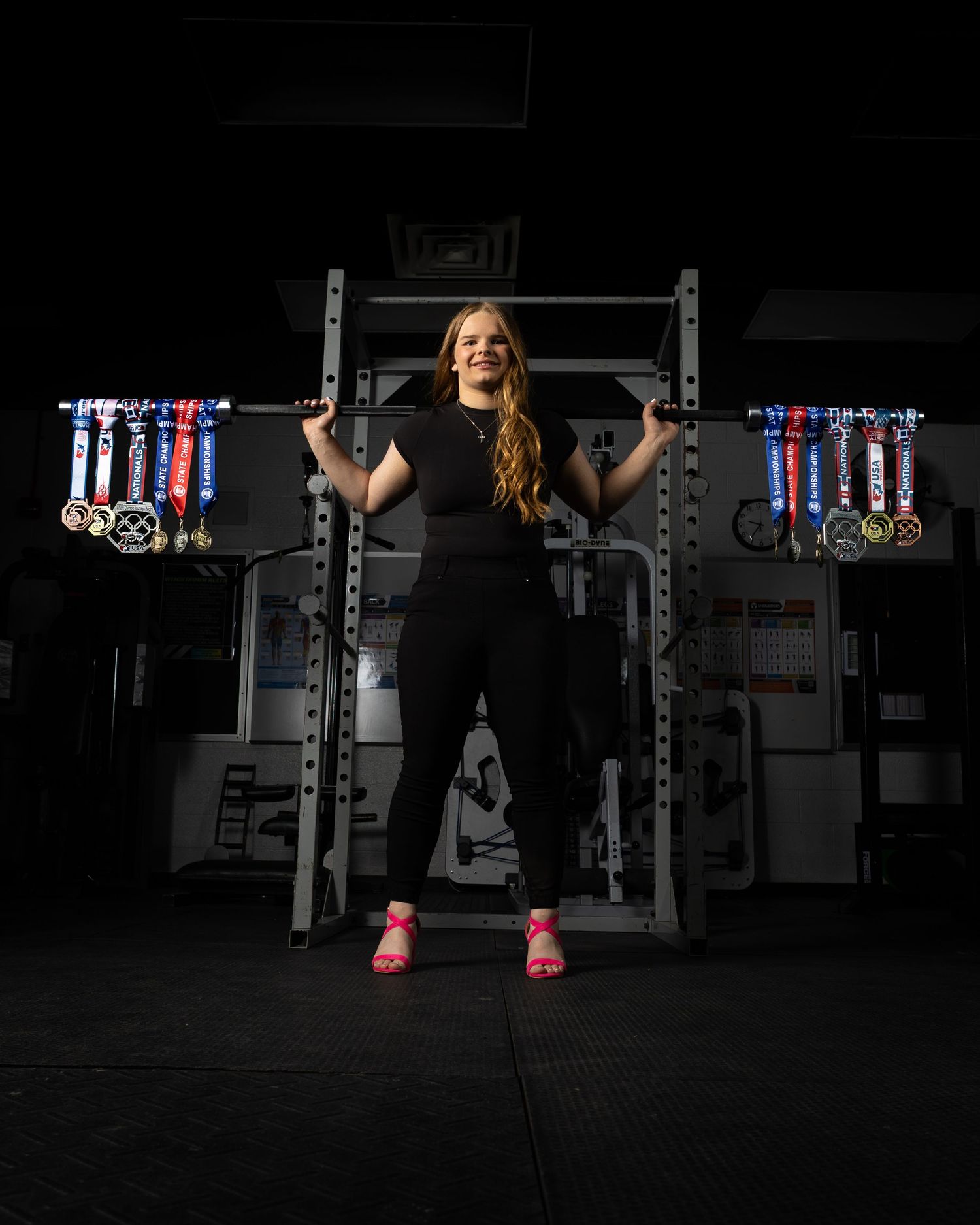 Young female athlete smiling in dark gym, holding barbell with colorful medals displayed on squat rack behind her.