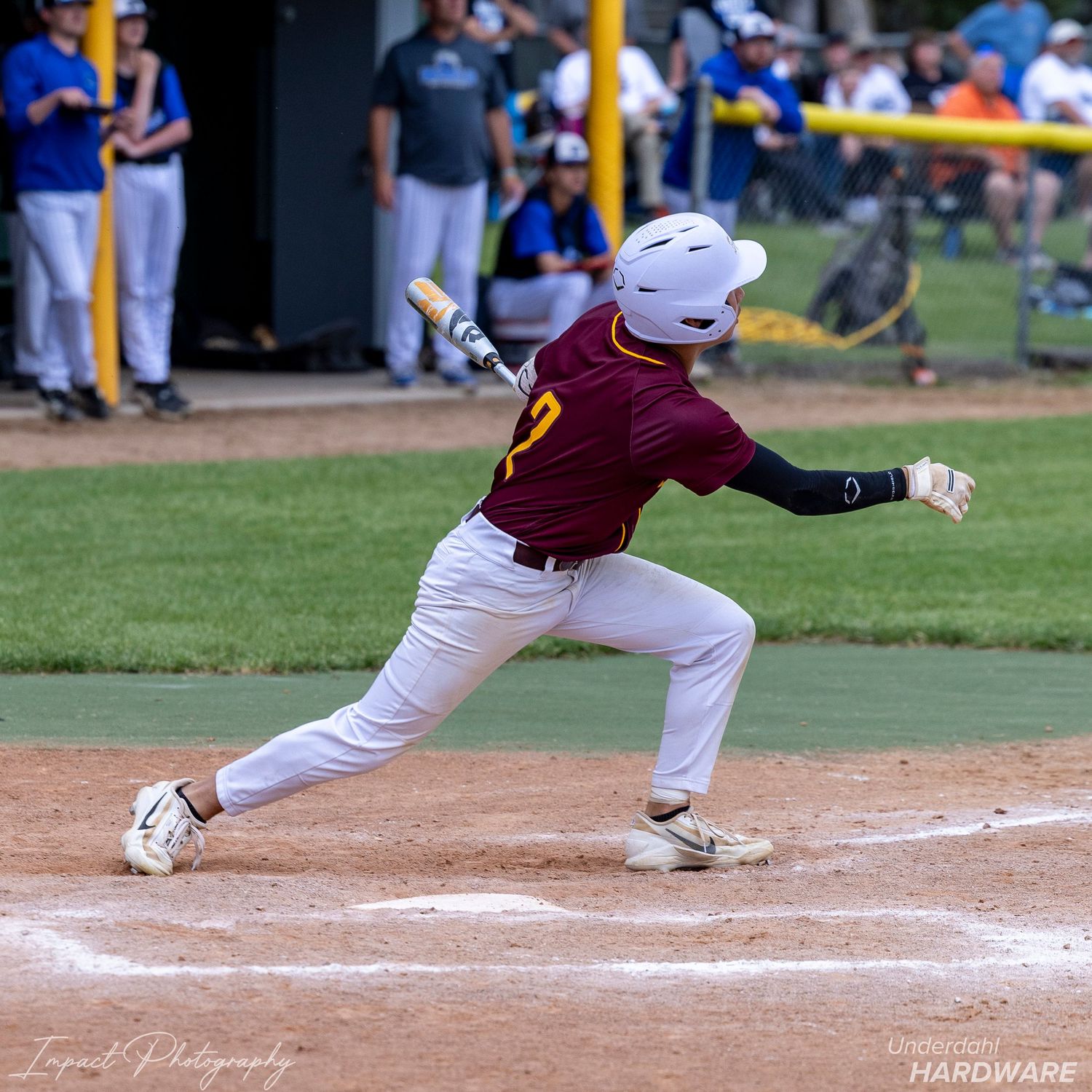 Youth baseball player in maroon uniform swinging bat at home plate during a sunny outdoor game.