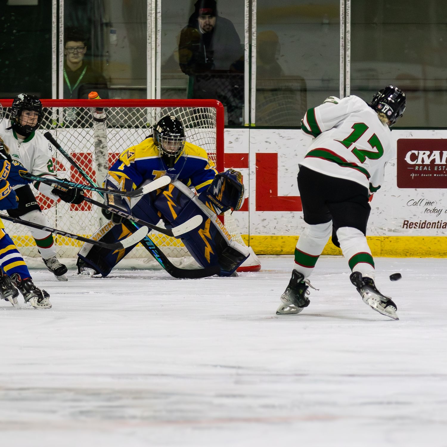 Hockey player #12 in white takes a shot on goal against blue and yellow goalie during an indoor ice hockey game.