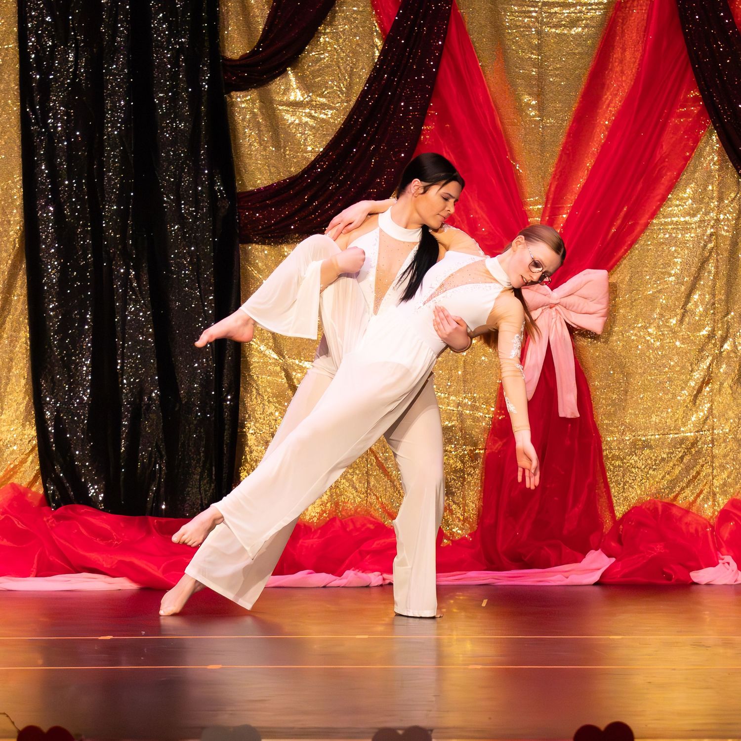 Two dancers perform a dramatic lift on stage in white costumes against a gold sequin backdrop.