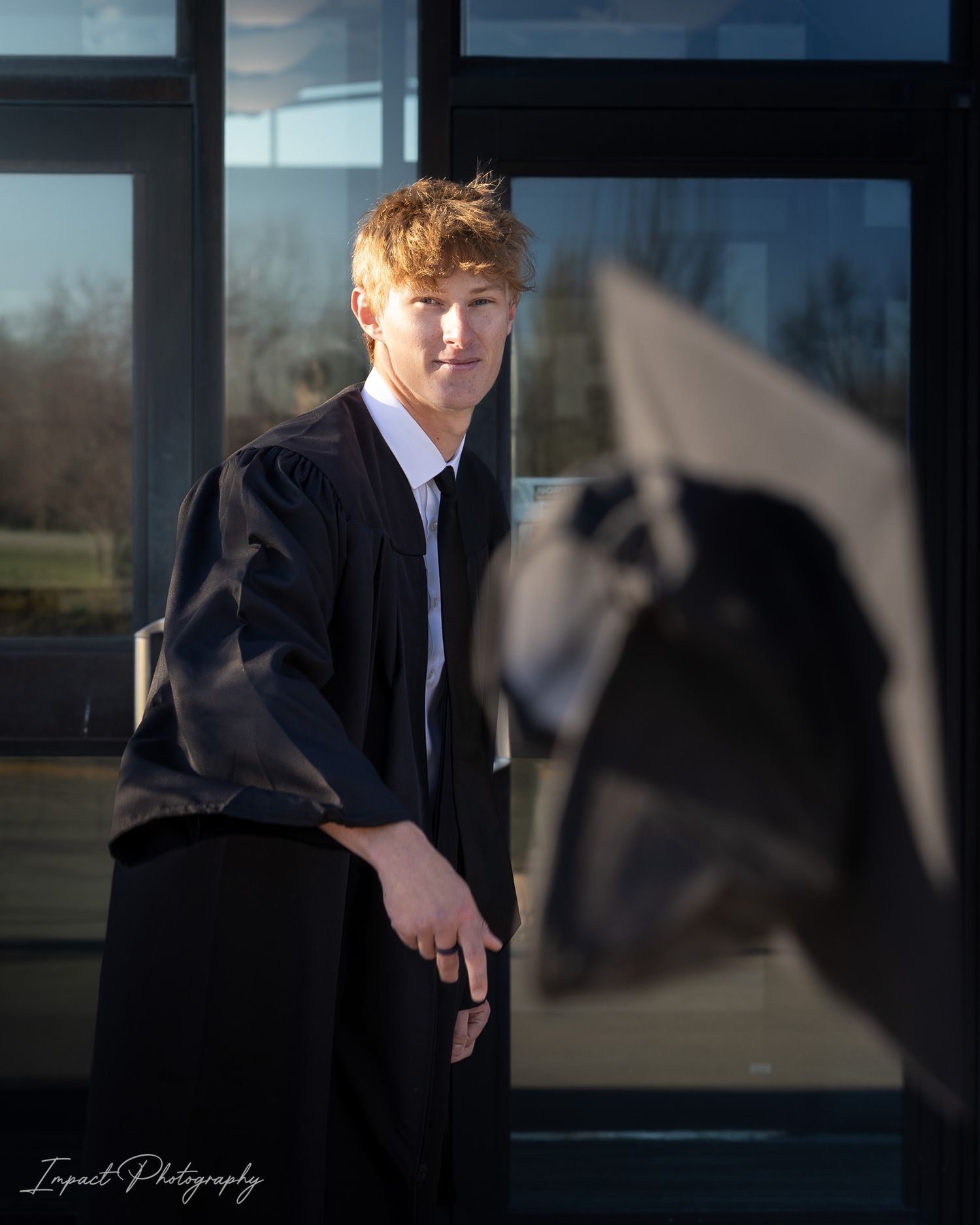 Young man in black graduation gown posing outdoors with a mortarboard cap in the foreground.