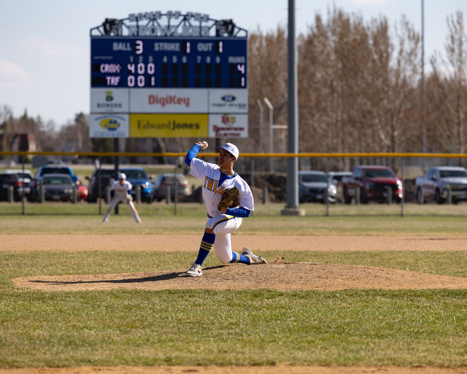 Baseball pitcher in blue uniform mid-throw on mound with scoreboard and sponsor signs visible in background.