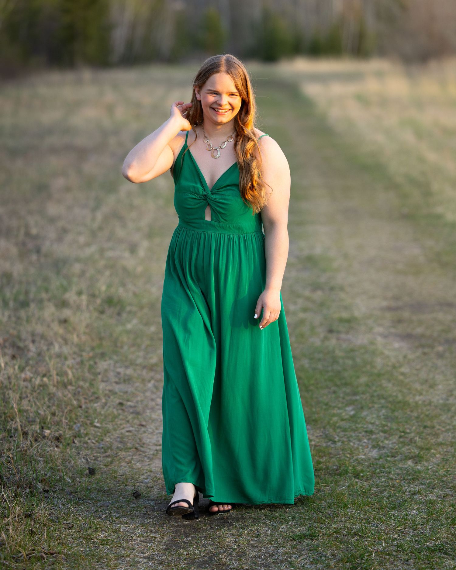 Young woman in elegant green maxi dress posing on a path in a field during golden hour.