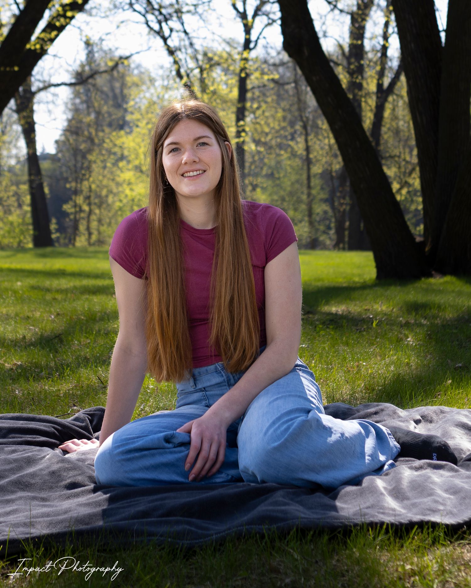 Young woman with long red hair sits on a blanket in a lush green park, smiling, wearing a maroon top and jeans.