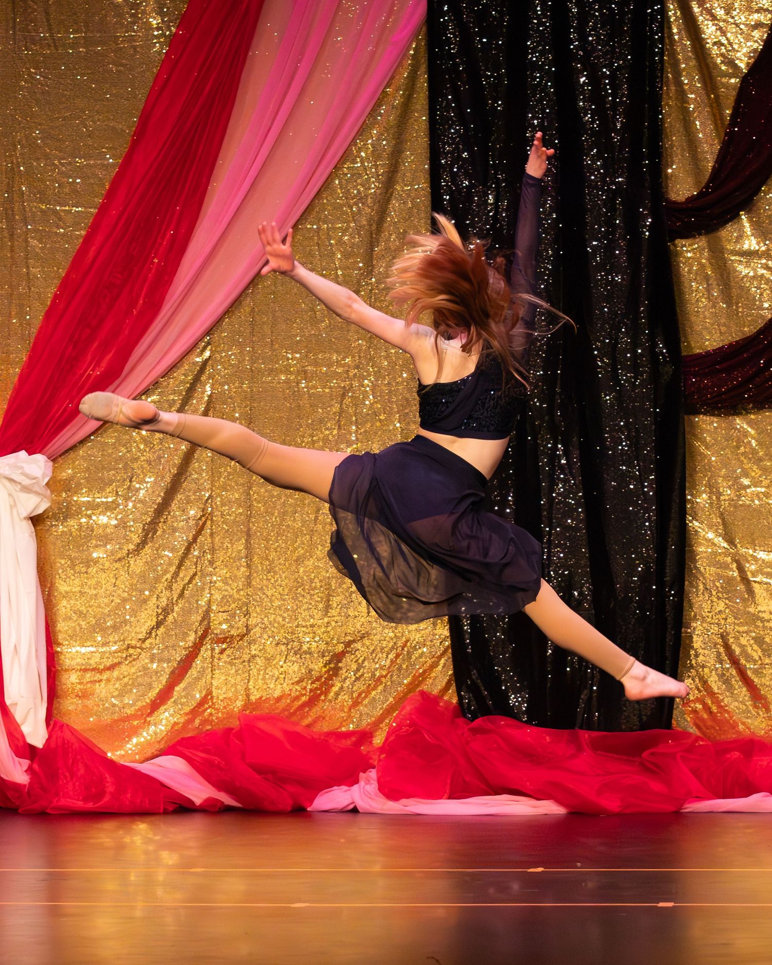 A young dancer leaps gracefully on stage in front of colorful gold and red sequined curtains.