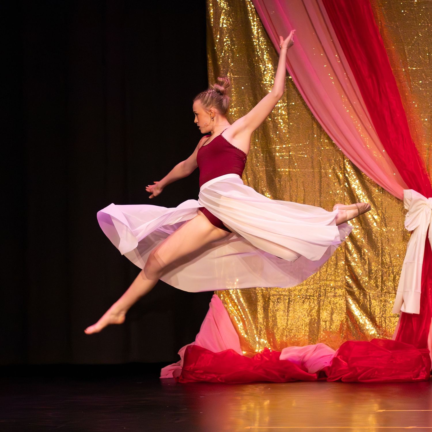 Young female dancer leaping gracefully on stage in a red and white flowing costume.