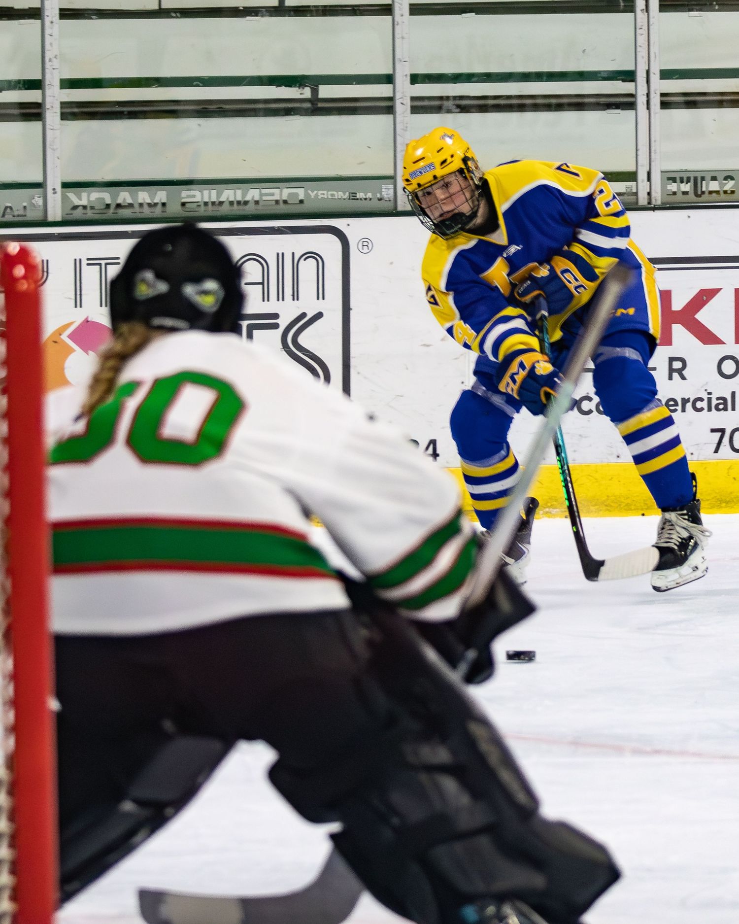 Blue and yellow hockey player winds up for a shot on goal against white-jersied goalie number 00.