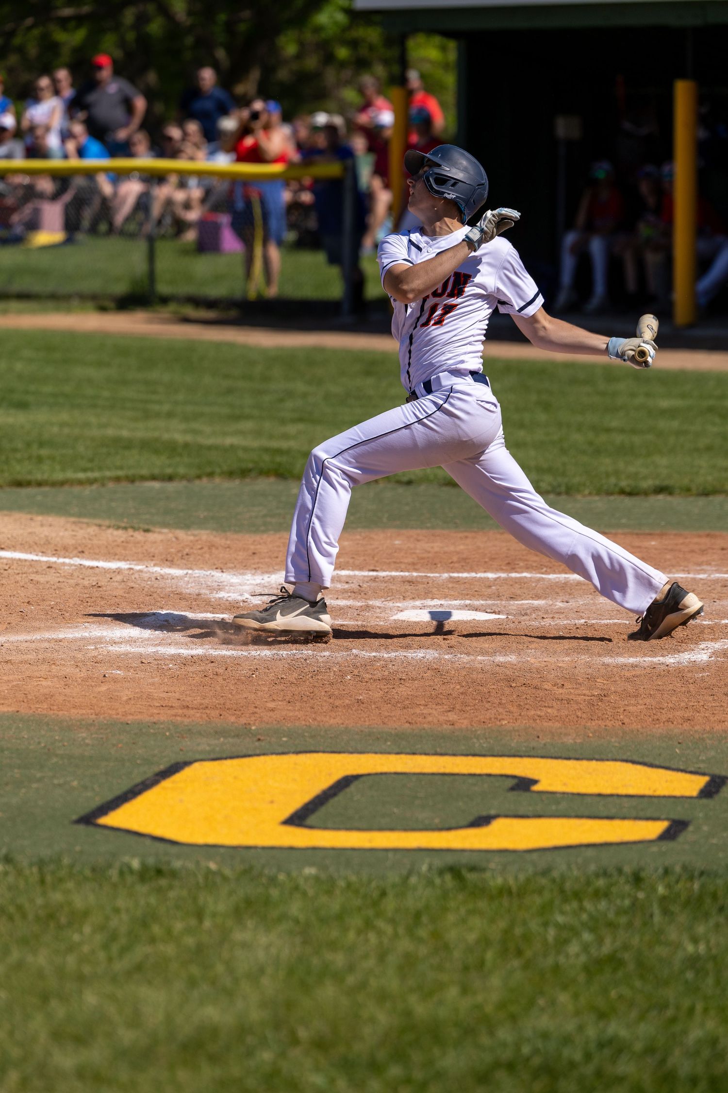 Baseball player in white uniform swings bat at home plate during a sunny outdoor game with crowd in background.