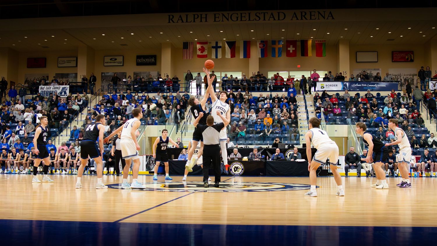 Basketball game tip-off at Ralph Engelstad Arena with players jumping for the ball before a packed crowd.